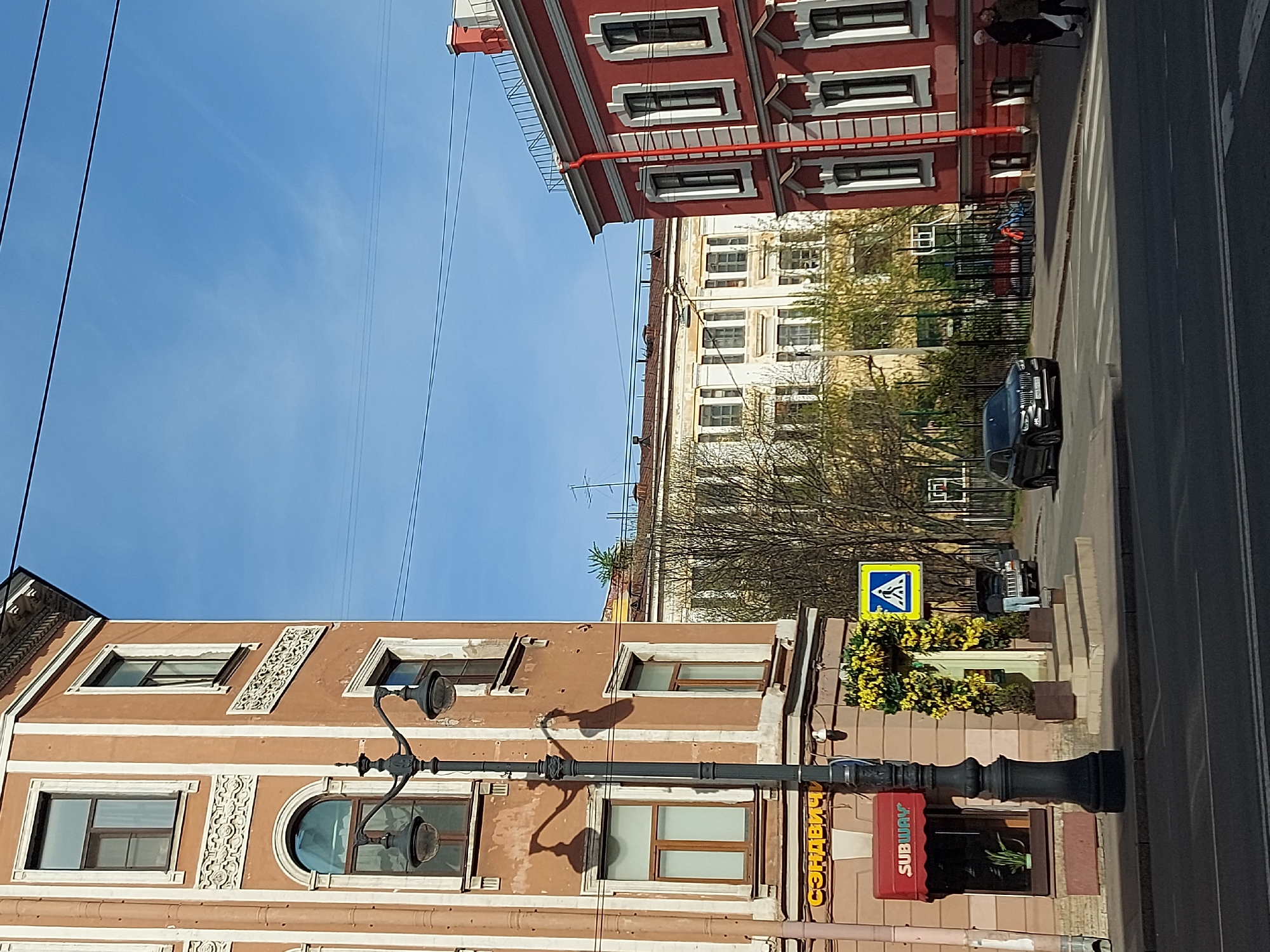 The image shows a street scene in Saint Petersburg, Russia. The street is lined with buildings of varying heights and architectural styles, featuring a mix of brown, red, and white facades with decorative elements. A pedestrian crossing sign is visible on the right side of the street, near a lamppost and a Subway restaurant sign. A black car is driving on the right side of the street. The sky is blue with powerlines overhead. The scene appears to be during the day with clear weather.