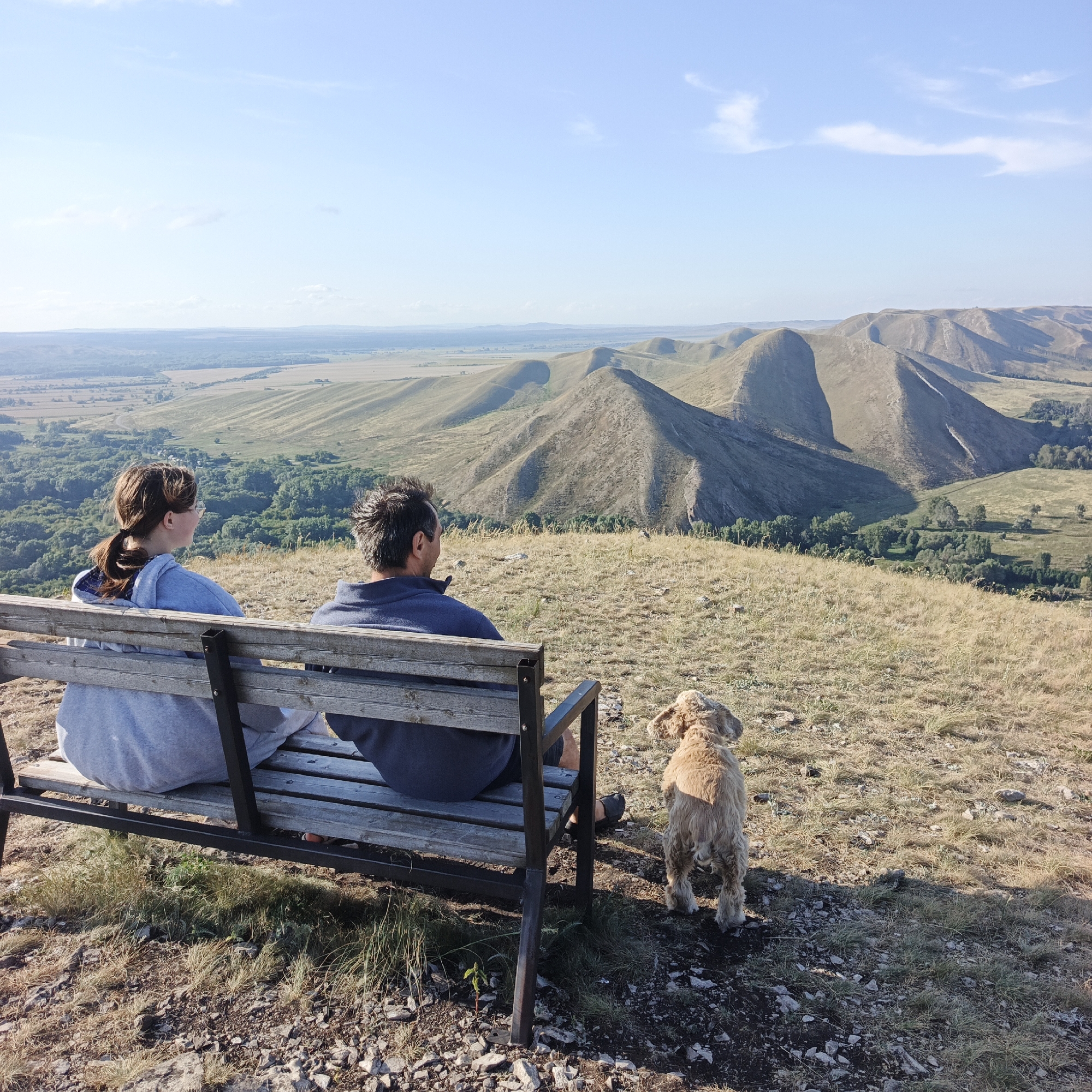 In the city of Andreevka, Russia, two people and a dog are situated on a wooden bench overlooking a vast, rolling landscape of hills under a clear blue sky. The man, on the right, wears a dark blue sweatshirt, while the woman, on the left, is in a grey hooded sweatshirt. The dog, a light brown spaniel-type breed, stands to the right of the bench. The bench itself is weathered wood with a metal frame and is positioned on a grassy, rocky incline. The background features a valley with trees and fields, leading to a distant mountain range with a textured, layered appearance. The sun casts long shadows, indicating it is either early morning or late afternoon.