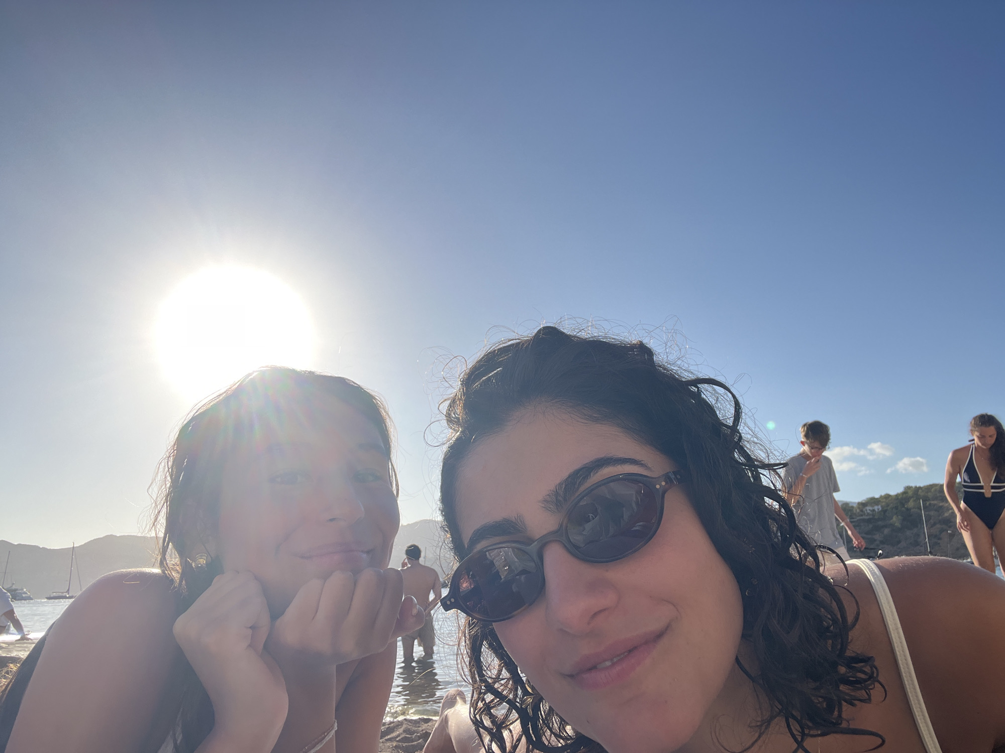 Two young women are lying on a sandy beach, facing the camera. The woman on the right is wearing sunglasses and has curly dark hair. The woman on the left has lighter brown hair and is resting her chin on her hands. Both appear to be sunbathing. In the background, there are other people in the water and on the shore. Several sailboats are visible in the bay. The sun is shining brightly, creating a lens flare in the upper left quadrant of the image. The environment is a beach with clear blue sky and calm water, indicative of a Mediterranean coastal location, likely Sant Josep de sa Talaia, Spain, known for its beaches and bays.