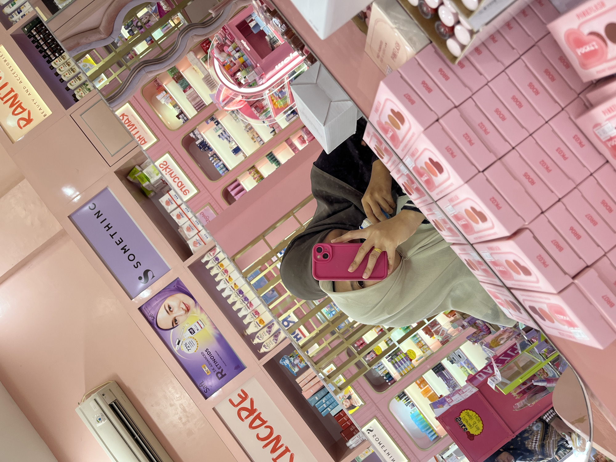 A woman wearing a beige hijab and a dark headscarf is taking a mirror selfie in a brightly lit, pink-themed cosmetics store. She is holding up a pink smartphone in her right hand, obscuring part of her face. Her left hand, adorned with a gold ring, is visible and holding the phone. The store's interior, reflected in a large mirror in the foreground, is filled with shelves displaying various beauty products, mostly packaged in pink and white. Notable signage includes "SKINCARE" and "SOMETHINC." In the background, a shelf displays bottles of "Safi 3X Advanced Retinoids," with a promotional image of a woman wearing a purple hijab. Another sign reads "RANITA BEAUTY AND ACCESSORIES." The overall impression is of a modern retail environment specializing in cosmetics, with a predominantly feminine aesthetic. The specific location is implied to be Lhokseumawe, Indonesia, based on the context provided.