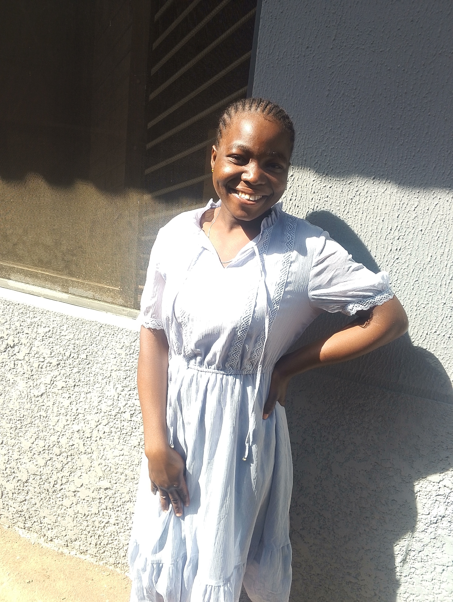The image captures a young Black woman smiling warmly at the camera, positioned outdoors in what appears to be Jos, Nigeria. She stands against a textured, light-colored wall, with a window featuring a dark screen or grille visible to her left. Her hair is styled in neat, dark cornrows.
The woman is wearing a light blue, short-sleeved dress that extends at least to knee-length. The dress has a V-neckline, a cinched waist, and a flowing skirt, adorned with white lace details on the bodice and sleeve hems. She poses with her right hand gently placed on her hip. The bright sunlight, indicative of a clear, sunny day, casts strong, defined shadows on the wall to her right and partially on her person, suggesting it is midday or early afternoon. A small, golden earring is visible on her left ear. The overall atmosphere conveyed is joyful and bright. There is no visible text in the media file.
