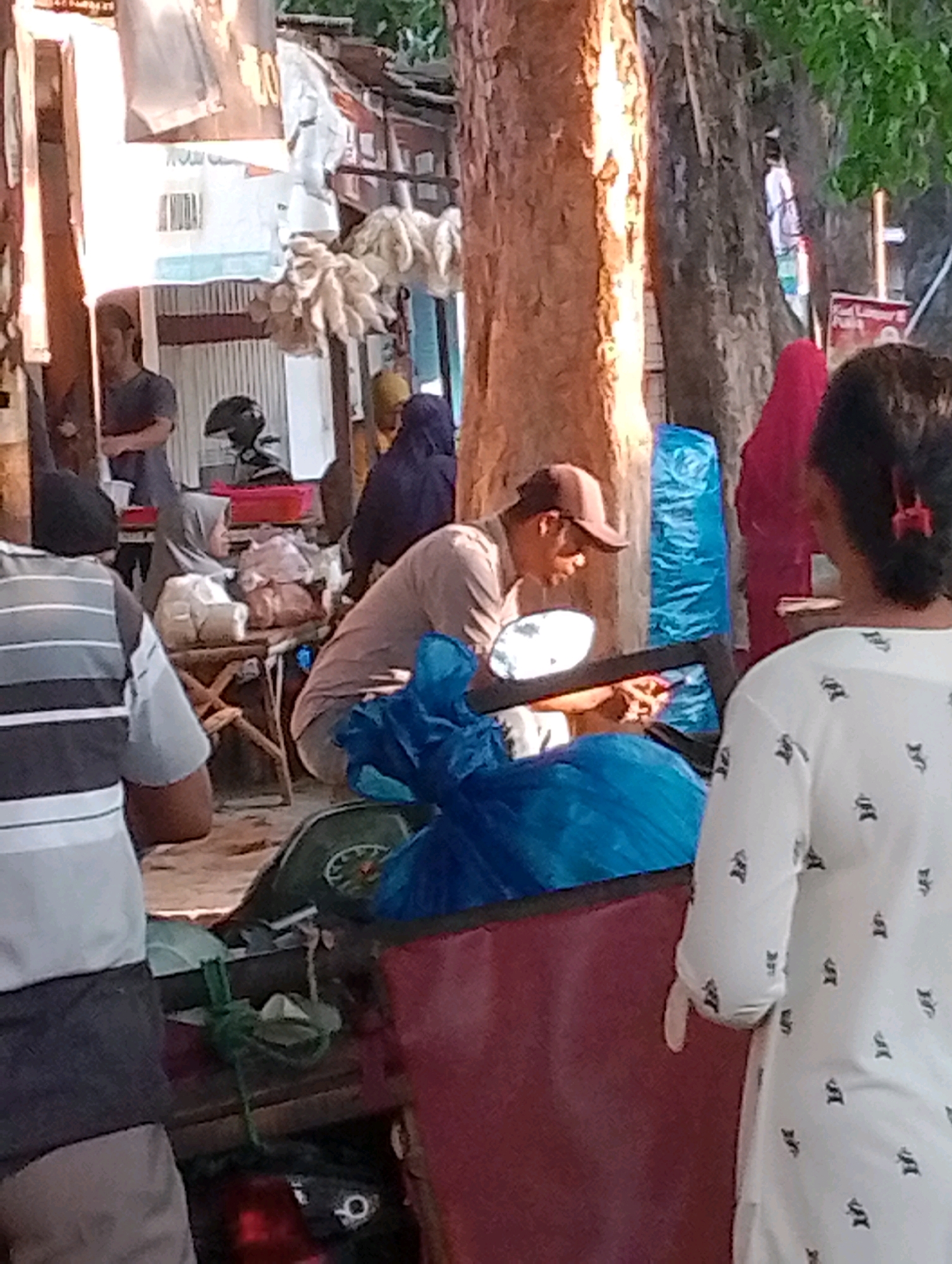 The image depicts a busy street scene, likely in Probolinggo, Indonesia, with several individuals and market stalls. In the foreground, a person wearing a grey and white striped shirt and a dark cap is partially visible, facing away from the camera. To their left, a person in a dark t-shirt and a cap is standing near a stall with various goods, including what appear to be bags of food items and some sort of white bundles hanging. To the right of the central figure, a man wearing a brown cap and a light-colored shirt is seated and appears to be working with something in his hands. He is situated next to a motorcycle mirror and several blue plastic bags. Behind him, a woman in a bright pink headscarf is partially visible. Further in the background, a woman in a dark blue headscarf is standing near some market stalls. The environment suggests a roadside market with temporary structures and a general atmosphere of commerce. The overall lighting indicates it is daytime.