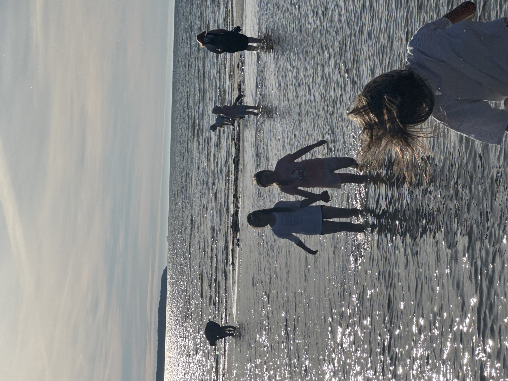 The image shows a group of people wading in shallow water, likely a beach or shoreline. The water is shimmering with sunlight, indicating it is daytime, possibly late afternoon due to the low angle of the sun and the long shadows cast. The sky appears bright and overcast with streaks of clouds. The people are scattered across the frame, with some closer to the foreground and others further back.
In the foreground, the back of a person wearing a white garment is visible, with their head and shoulders angled downwards as if interacting with the water. To their left, another person is partially visible, appearing to be in a similar pose. Further back, a child in a light-colored t-shirt and dark shorts is reaching out their arms. Behind them, another child or young person in a darker t-shirt and shorts is bending over. In the mid-ground, a person in a dark dress is standing. Another figure is visible further back, also standing in the water. The water is shallow and appears to be a muddy or sandy bottom, with ripples and reflections from the sunlight. The overall mood is one of casual recreation and enjoyment of the water. There are no visible objects other than the people themselves and the natural elements of water and sky. The setting suggests a temperate climate.