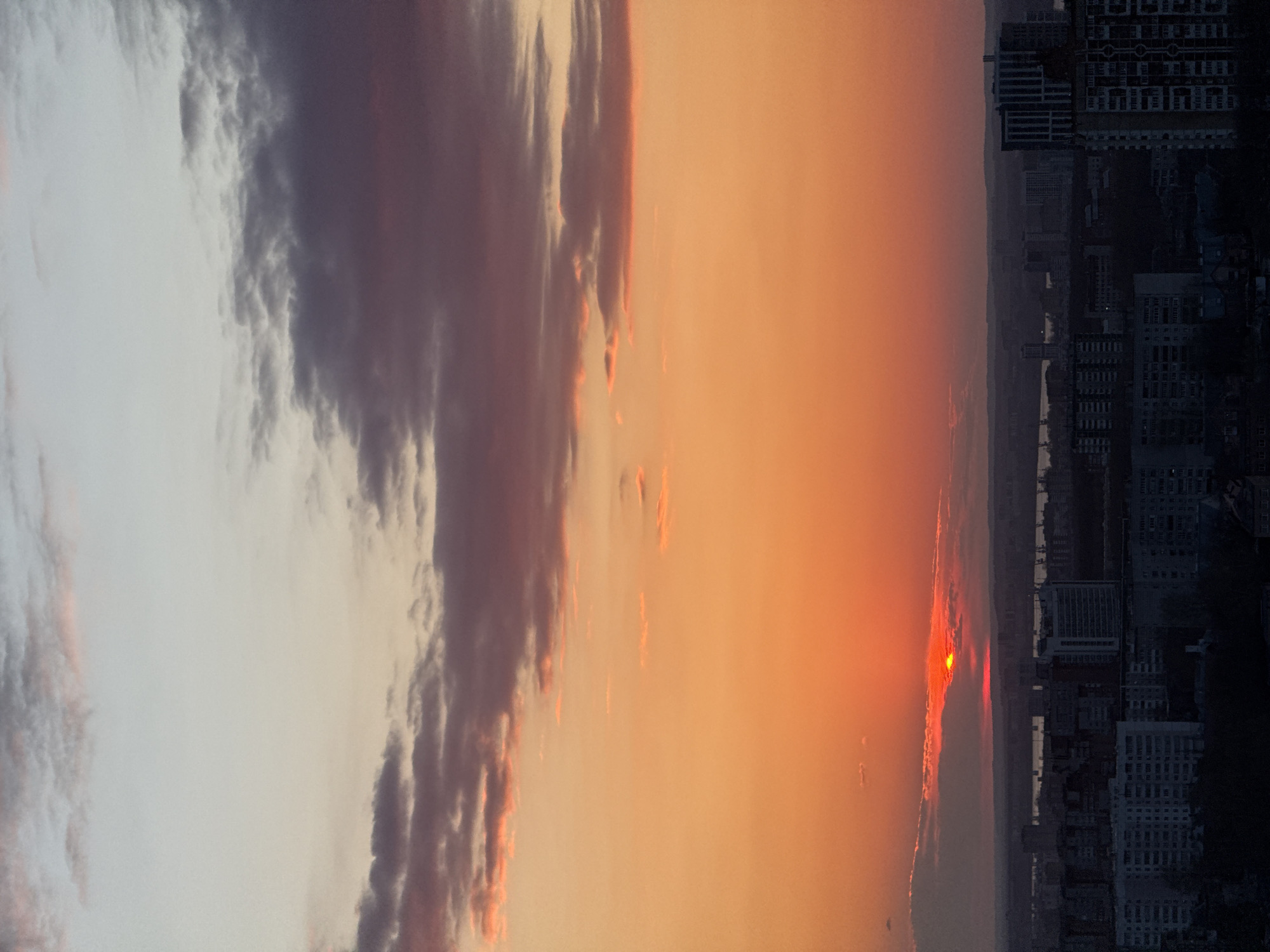 This panoramic image captures a dramatic sunset or sunrise over a city skyline, likely Perm, Russia. The sky dominates the frame, bursting with vibrant hues of orange and red near the horizon, gradually transitioning to lighter yellows, grays, and purples higher up. The sun, a brilliant orange-red orb, is visible low on the horizon, partially obscured by a narrow band of clouds or atmospheric haze, casting an intense glow across the clouds directly above and around it.
To the left, a large, dense formation of dark, voluminous clouds stretches across the sky, displaying deep gray and purple tones. The edges of these clouds and some smaller, more fragmented clouds closer to the sun are illuminated with striking orange and pink highlights from the low-angle light.
Beneath the colorful sky, the distinct silhouette of a city stretches across the horizon. Numerous buildings of varying heights and architectural styles are visible in dark relief against the bright sky, forming a dense urban landscape. A faint line of water, possibly a river like the Kama, appears to weave through or past the city, reflecting some of the sky's light. Distant hills or land formations are also discernible as subtle, darker layers beyond the immediate urban sprawl.
The image conveys a sense of peaceful grandeur, capturing the natural beauty of a dawn or dusk sky over a bustling city. No people or specific activities are visible, and there is no discernible text. The scene is characterized by its powerful color palette and the serene contrast between the dynamic sky and the static urban environment.