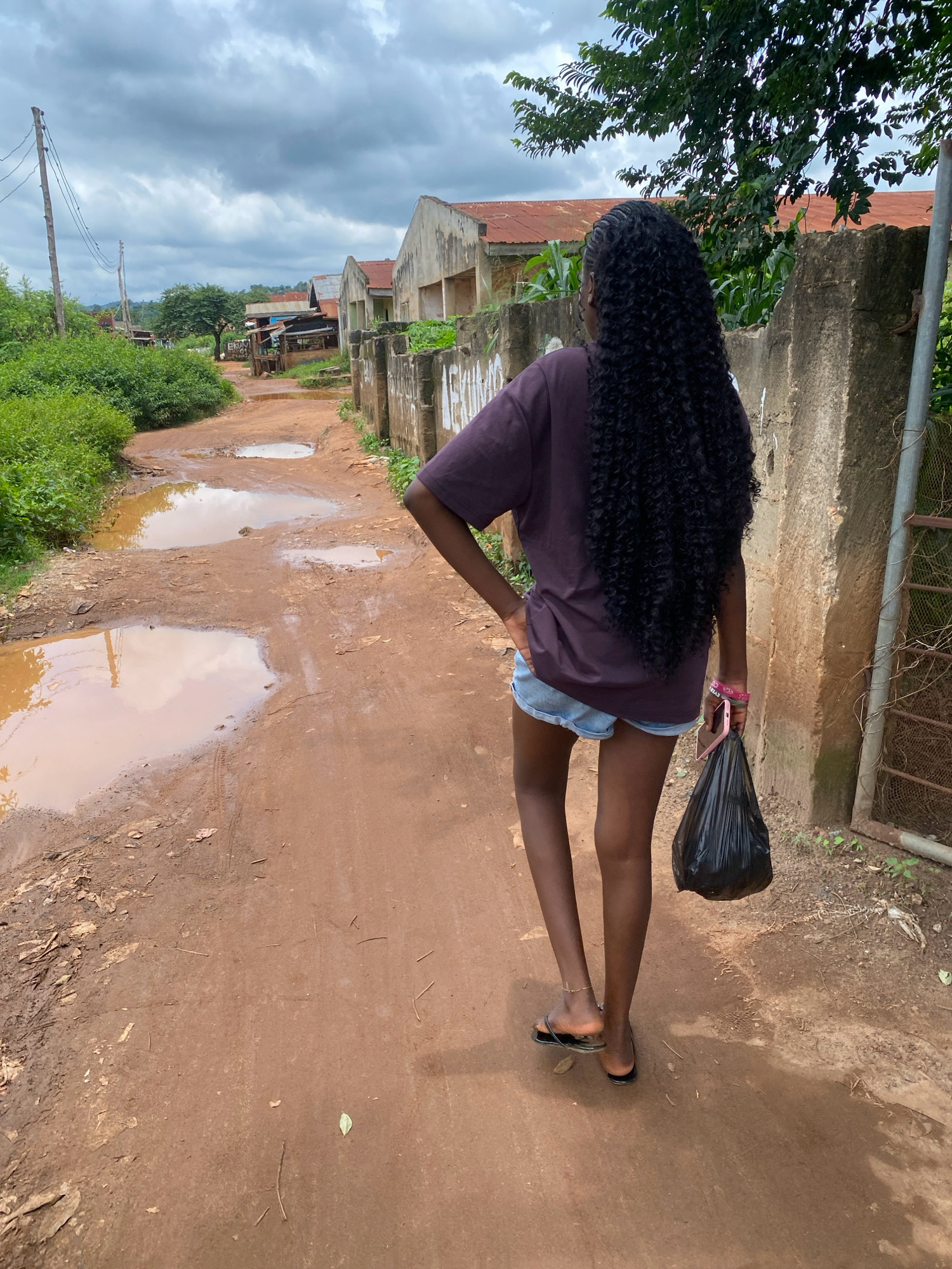 A young Black woman walks away from the camera down a muddy, unpaved road in Iworoko, Nigeria. She has long, dark, curly hair and wears a dark purple t-shirt and denim shorts. She carries a black plastic bag in her right hand and wears black flip-flops. The road is lined with puddles of water. On the right side of the road, there is a concrete wall and a metal fence. Behind the wall, there are buildings with corrugated metal roofs and overgrown vegetation. The sky is cloudy and gray.