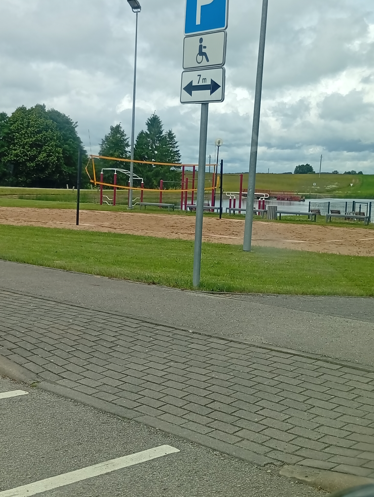 The image shows a disabled parking sign with a distance indicator for 7 meters, located in Šaukėnai, Lithuania. The sign is positioned next to a volleyball court with a sand surface and a net. In the background, there is a body of water, grassy hills, and trees under a cloudy sky. A paved pathway with a concrete curb runs in the foreground.