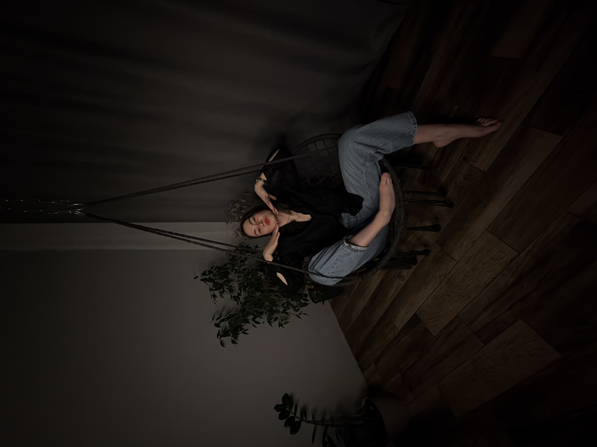 The image captures a relaxed, intimate indoor scene, featuring a young woman seated in a dark, round hanging chair. She is positioned horizontally within the frame, suggesting the photo was taken with the camera rotated. The woman has dark hair and wears a black top and light blue jeans, with bare feet. She has red lipstick and gazes towards the viewer with a serene or calm expression, one hand resting near her chin and the other on her torso.
The setting appears to be a dimly lit room, likely a home, with a dark wooden floor visible on the right side and below the chair. The background is a plain, dark grey or black wall, contributing to the subdued atmosphere. Two potted plants add a touch of greenery to the scene; a larger, dense-foliaged plant is placed behind the chair, and a smaller plant with darker leaves is visible in the bottom left corner.
The lighting is very low, creating deep shadows and emphasizing the glow of the woman's face and hands. The time of day is not explicitly clear but the dim illumination suggests either evening or a room with minimal natural light. There is no visible text in the image. The overall mood is quiet and contemplative, portraying a moment of repose in Chaikovsky, Russia.
