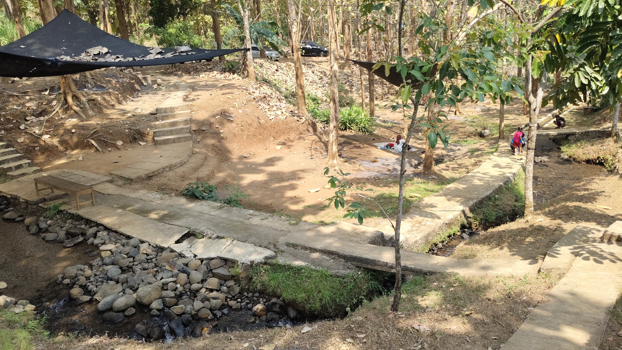 The image depicts an outdoor scene in a wooded area, possibly a park or natural reserve, located in Kresek, Indonesia. In the foreground, a narrow stream flows over a bed of rounded rocks. A concrete pathway runs alongside the stream, with another concrete bridge spanning it further back. On the left bank of the stream, a wooden bench sits on a concrete platform. Behind this, a concrete staircase leads up a slope.
Further into the scene, two cars are parked on a dirt area. Several black tarp structures are visible, providing shade in different parts of the area. A group of people are present: two individuals, one in a pink shirt and the other in a blue shirt, are walking on the concrete path. Another person is sitting further back on the path, and a fourth individual is seated on a mat or blanket on the ground under a tree. The environment is characterized by numerous trees with bare branches, dry leaves scattered on the ground, and patches of green vegetation. The overall impression is a relaxed, natural setting with amenities for visitors.