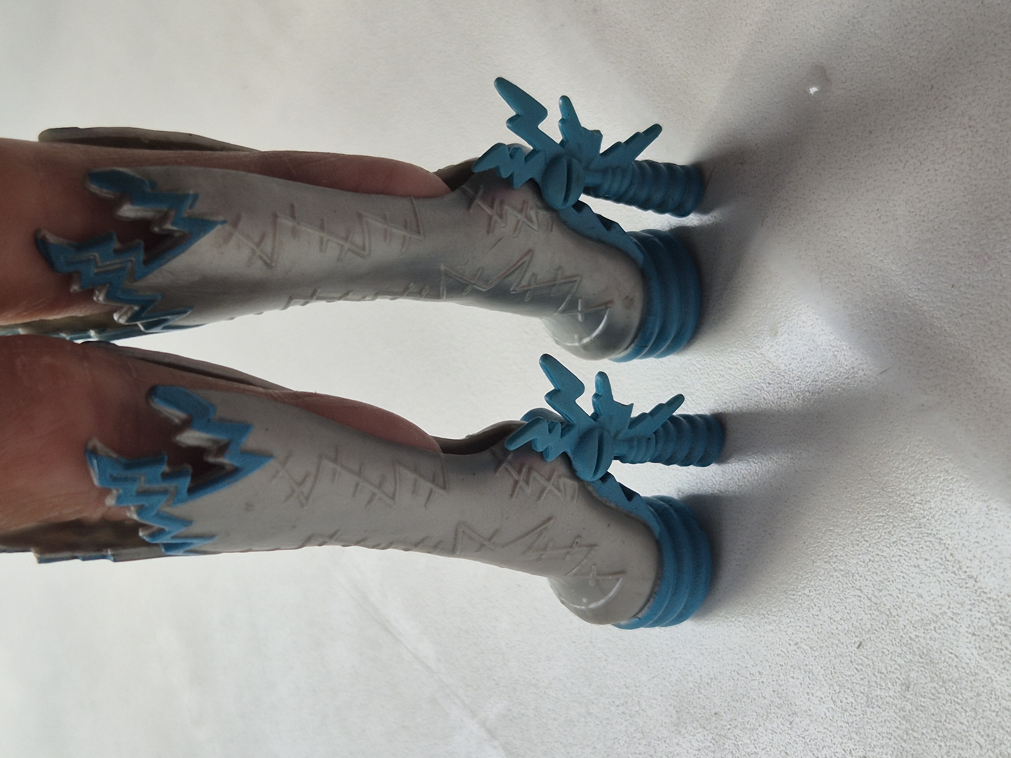 The image shows a close-up, overhead view of two toy dinosaur feet. The feet are predominantly a light gray color with jagged, turquoise accents resembling lightning bolts or flames around the ankles. The soles of the feet are a solid turquoise and appear to have a ribbed texture. Each foot also features a turquoise, three-pronged decorative element extending from the top of the ankle. The toy is held by a person's hands, which are visible on either side of the feet. The background is a textured, off-white surface, possibly a table or floor. The lighting suggests an indoor setting, as there is a shadow cast by the toy. No text is visible in the image.