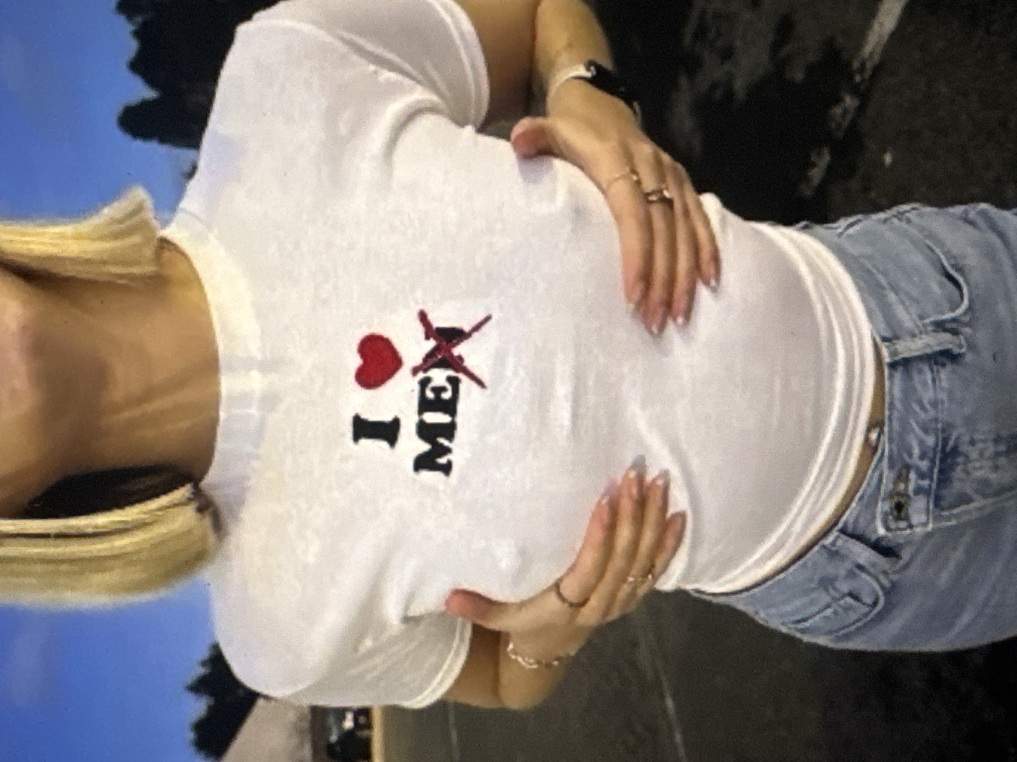 A person with blonde hair, wearing a white t-shirt with the text "I ❤️ ME" and a red X over the letter "M", and light blue jeans, stands in what appears to be an outdoor parking lot at dusk. The person's hands are positioned on their chest, with rings on their fingers. A bracelet is visible on their left wrist. The background shows trees and a dusky sky. The location is Kuta Selatan, Indonesia.