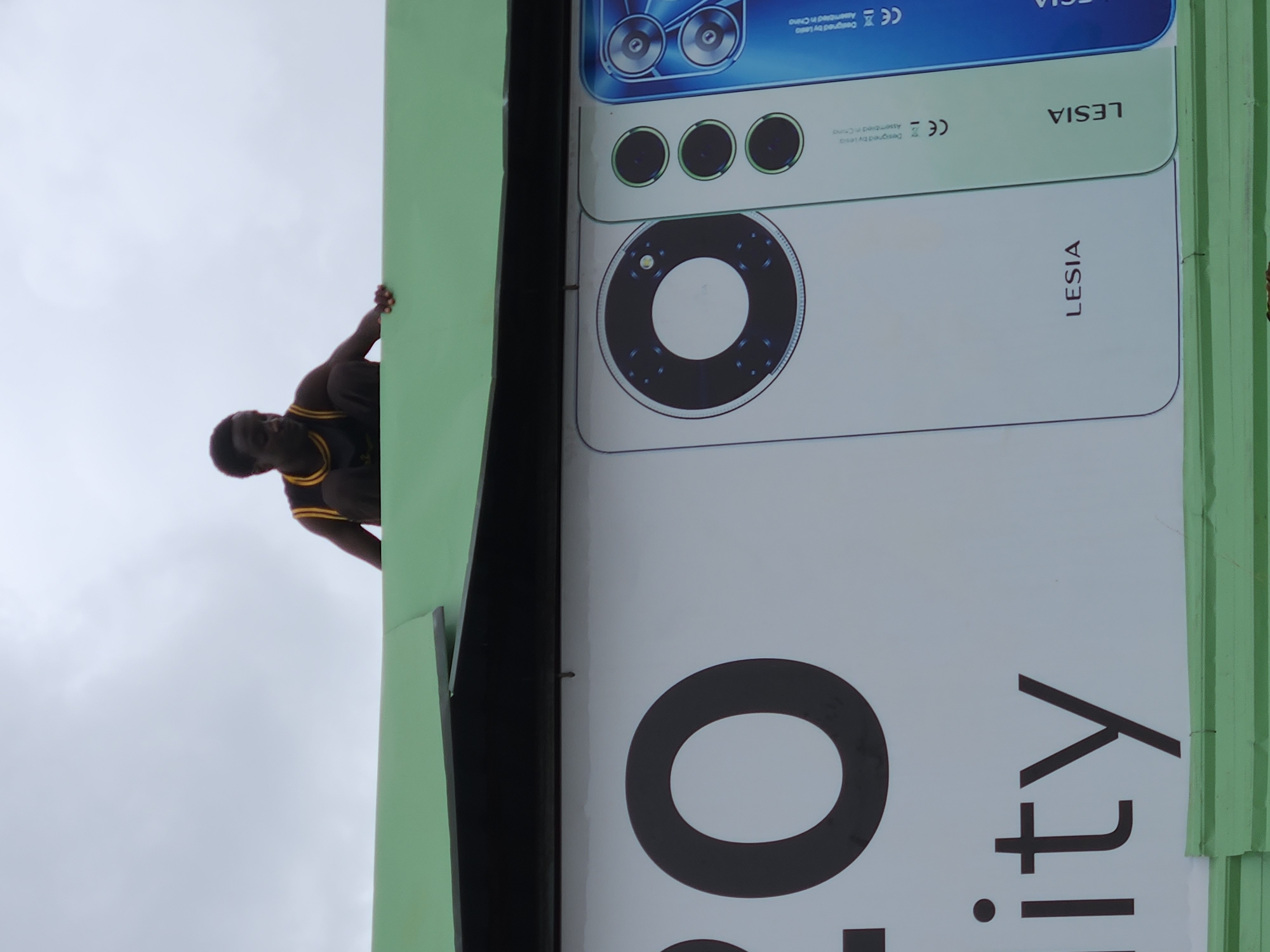 A dark-skinned person wearing a black tank top with yellow trim is crouched on top of a green corrugated metal structure, possibly a roof or awning. The person is looking down and to the left, with their right hand resting on the surface they are on. Below the person is a large advertisement featuring a white mobile phone with a circular camera module. The advertisement also displays large black text, including the number "20" and the word "ity". The background is an overcast sky.