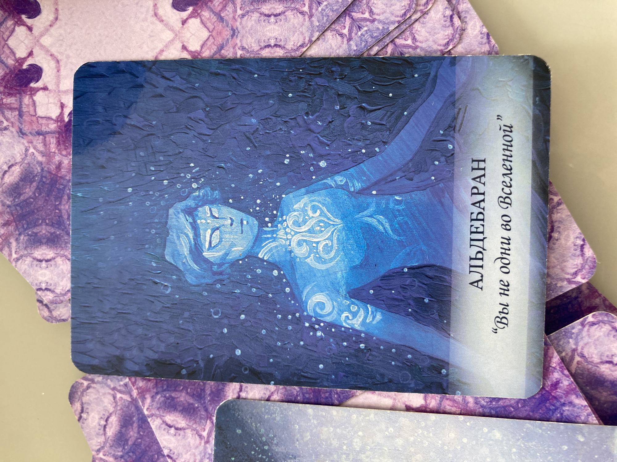 A tarot-style card titled "АЛЬДЕБАРАН" (Aldebaran) with the phrase "Вы не одни во Вселенной" ("You are not alone in the universe") is the primary subject of the image. The card features an illustration of a person rendered in blue tones, seemingly floating in a space filled with small white dots, reminiscent of stars. Decorative white swirls adorn the person's chest. The background is a deep blue, possibly representing the cosmos. The card is lying on top of other similar cards that have a purple and white watercolor-like pattern. No specific location in Florianópolis, Brazil, is identifiable from the visual information provided.
