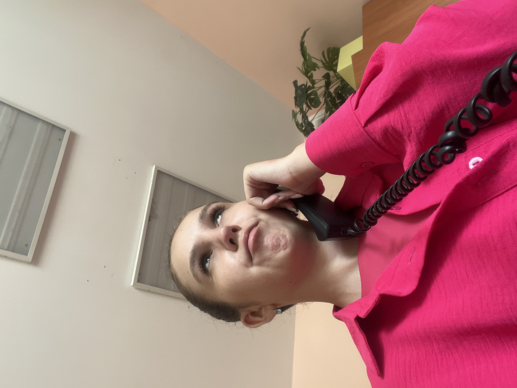 A young woman, wearing a bright pink collared shirt, holds a black landline telephone to her ear. She is looking up and to her left with a thoughtful or questioning expression. Her hair is pulled back in a bun. The background consists of a white ceiling with two recessed light fixtures, a peach-colored wall, and a portion of a wooden desk with a potted plant. The scene appears to be indoors, likely an office or home environment. There is no indication of the specific location within Novoaltaysk, Russia.