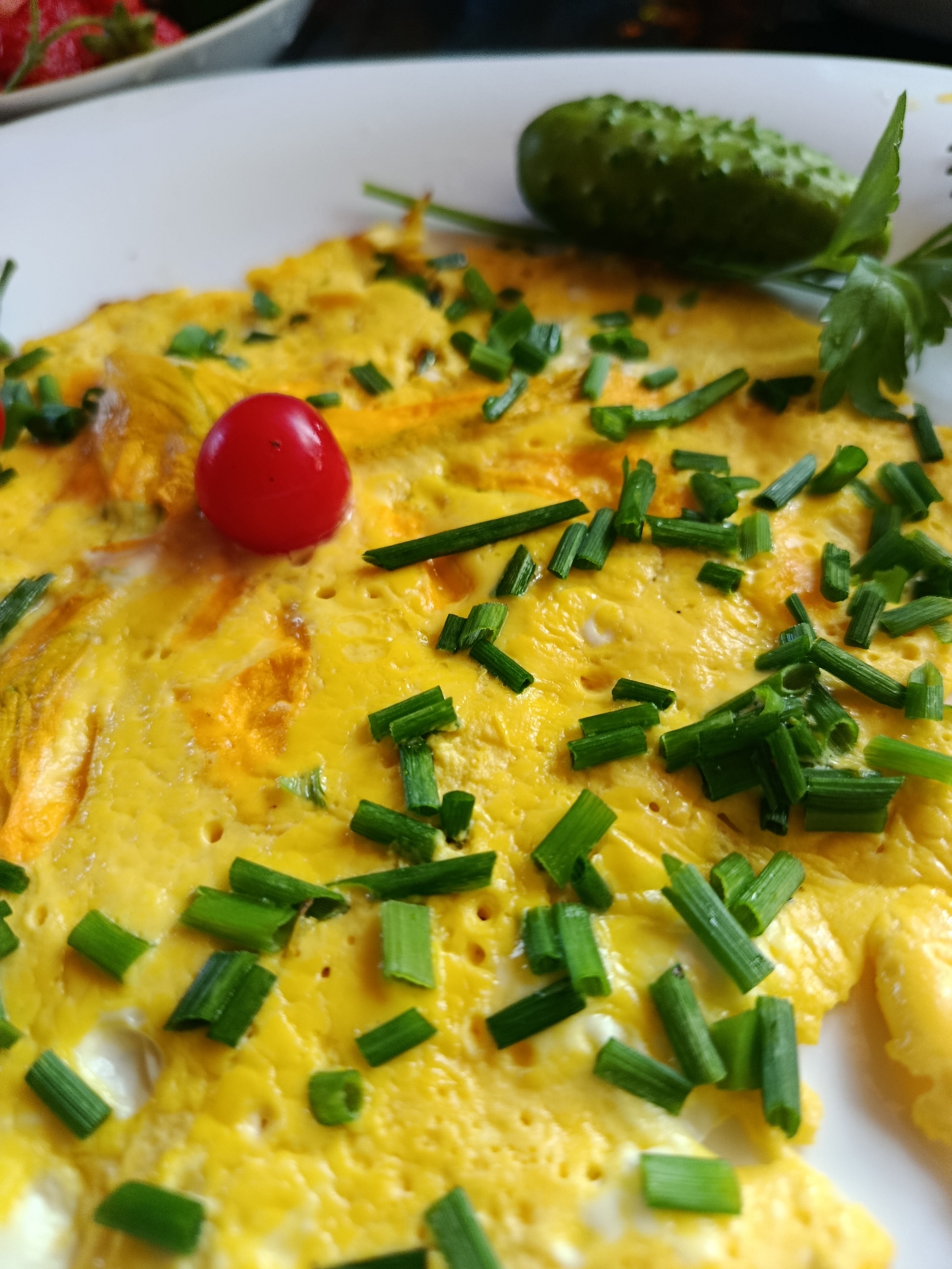An omelet topped with chopped chives and a single cherry tomato sits on a white plate. To the right of the omelet, a small pickled cucumber and a sprig of parsley are visible. In the upper left corner, a portion of a bowl filled with strawberries can be seen. The image does not contain information about the city of Верея, Russia, nor does it depict any people, actions, or interactions.