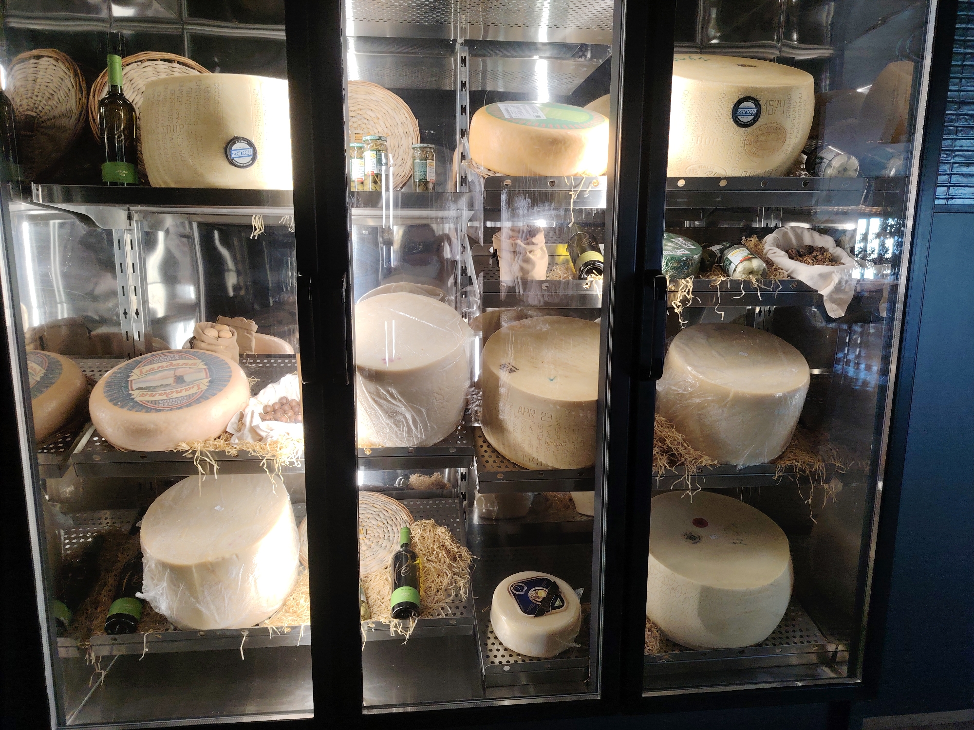 The image shows a refrigerated display case filled with wheels of cheese and a few bottles of wine. The display case is divided into multiple shelves, each holding different sizes and types of cheese. Some of the cheese wheels are very large, while others are smaller. Several of the cheese wheels have labels or stamps on them. There are also some woven baskets and straw-like material used for presentation. The lighting inside the display case is bright, highlighting the products. The exterior of the display case appears to be made of glass and metal, with black framing. The background of the image is blurred, but it suggests an indoor setting, possibly a market or specialty food store. There are no people visible in the image. The location is identified as Konigil, Uzbekistan.