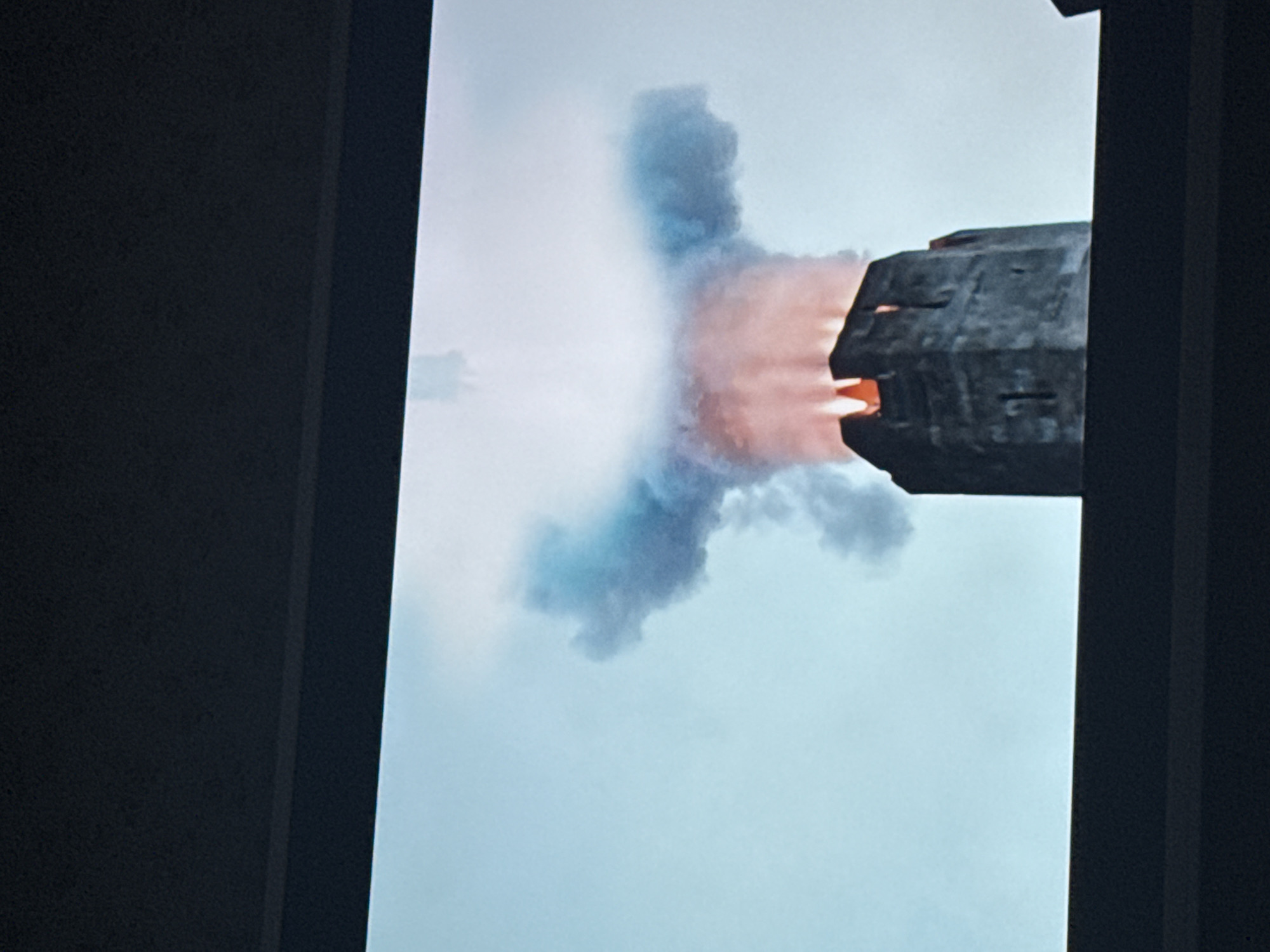 The image shows a spacecraft engine emitting exhaust. The exhaust is orange near the engine and fades to blue as it expands. The environment appears to be a cloudy or overcast sky. The spacecraft engine is a dark, blocky shape with visible details suggesting a complex structure. No location or people are visible in the image. There is nothing suggesting Mytishchi, Russia.