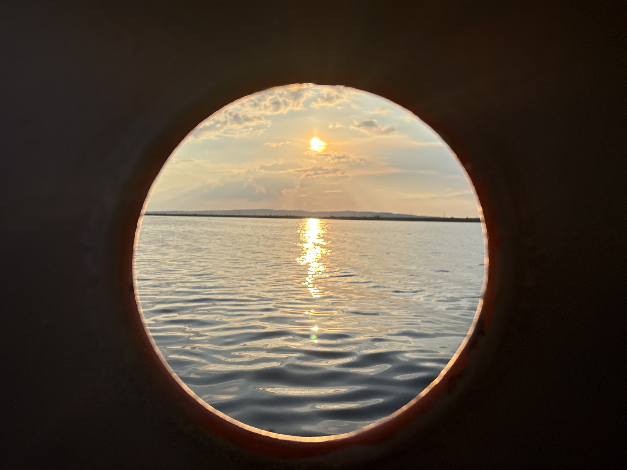 A circular frame, possibly a porthole, captures a sunset over a body of water. The sky is filled with soft clouds illuminated by the setting sun, which reflects a golden path across the rippling water. A distant horizon line separates the sky and water. The foreground is dominated by the dark, textured frame of the porthole, creating a sense of looking out onto the scene. The location is identified as Chaikovsky, Russia.