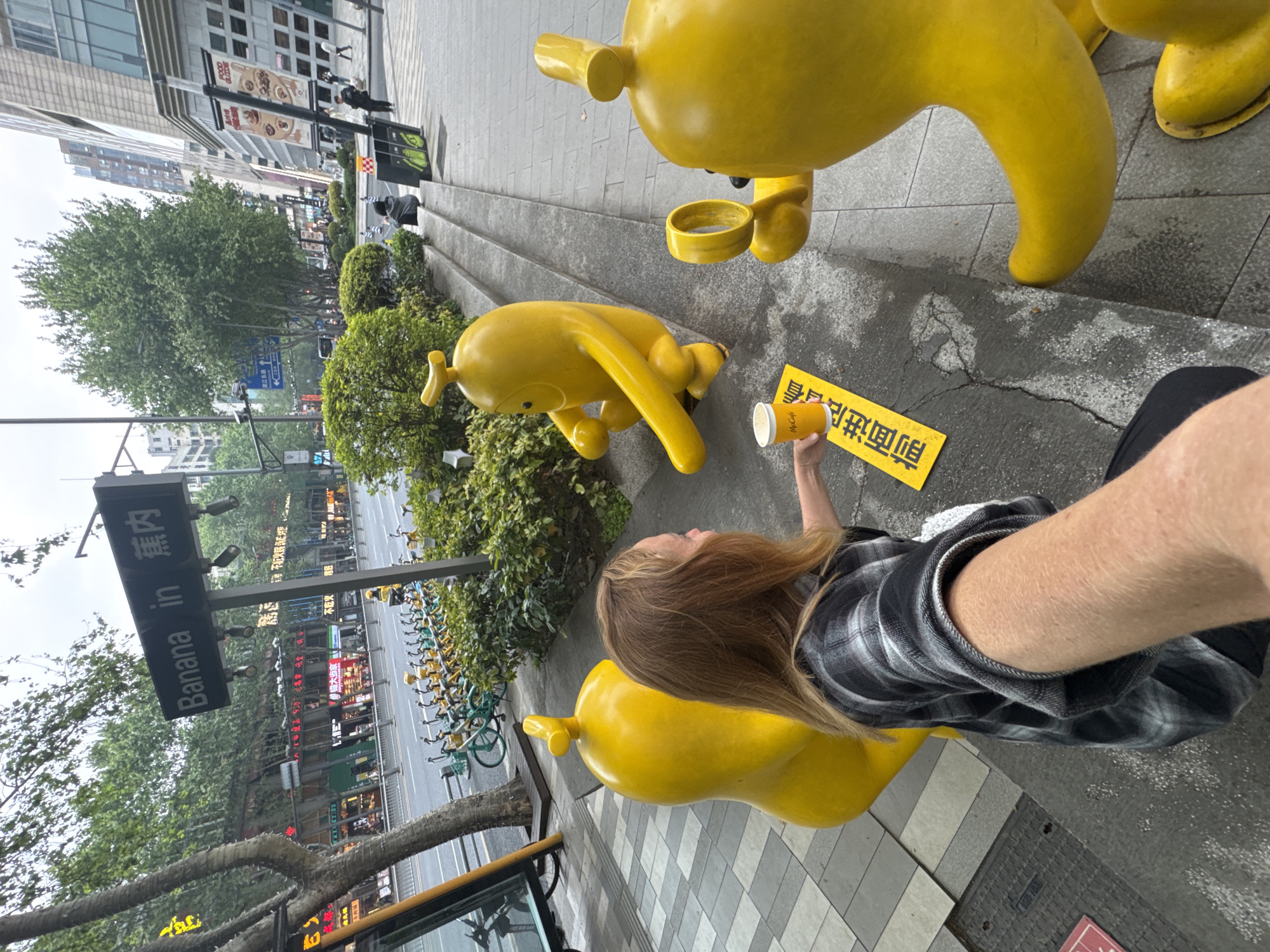 A low-angle, wide-angle shot depicts a street scene in Jinjiang District, China, dominated by yellow, stylized banana-shaped sculptures. A light-skinned person wearing a gray and black plaid shirt holds a cup bearing the word "McCafe." A sign with "Banana in" is also visible. Bicycles are parked in a row along the street. The buildings in the background suggest an urban environment.