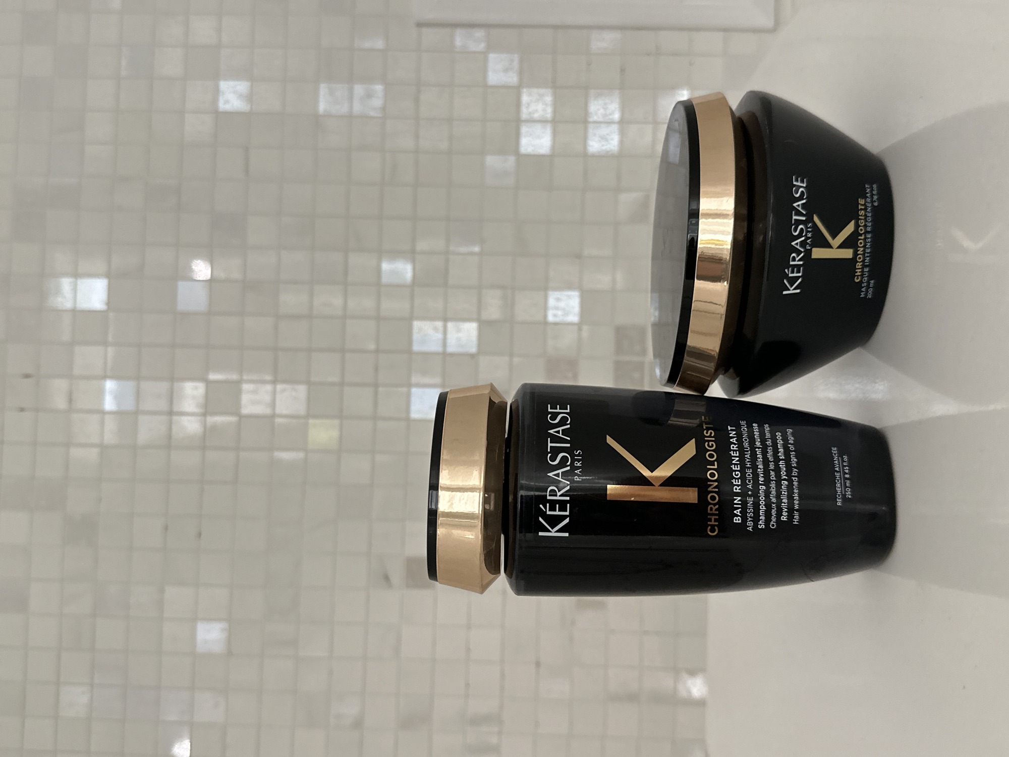 The scene is situated in Asenovgrad, Bulgaria. Two Kérastase Paris Chronologiste haircare products are positioned on a glossy white surface, against a background of light-colored square mosaic tiles.
On the left, a black bottle with a gold-colored cap features the Kérastase logo and "CHRONOLOGISTE" branding in white text, with a prominent gold "K". Text on the bottle identifies it as "BAIN RÉGÉNÉRANT" (Revitalizing youth shampoo) containing Abyssinia and Hyaluronic Acid, designed for hair weakened by signs of aging, with a volume of 250 ml (8.45 fl.oz.).
To the right, a black, broad-based jar with a gold-colored lid displays similar Kérastase Paris Chronologiste branding. This product is labeled "MASQUE INTENSE RÉGÉNÉRANT" (Intense revitalizing mask) and contains 200 ml (6.76 fl.oz.).
Both products exhibit subtle reflections from the ambient lighting on their surfaces. The glossy white surface beneath them also reflects their forms. The background wall consists of evenly spaced, reflective square tiles, which show varying degrees of light and shadow, indicating an indoor environment. No people are present.