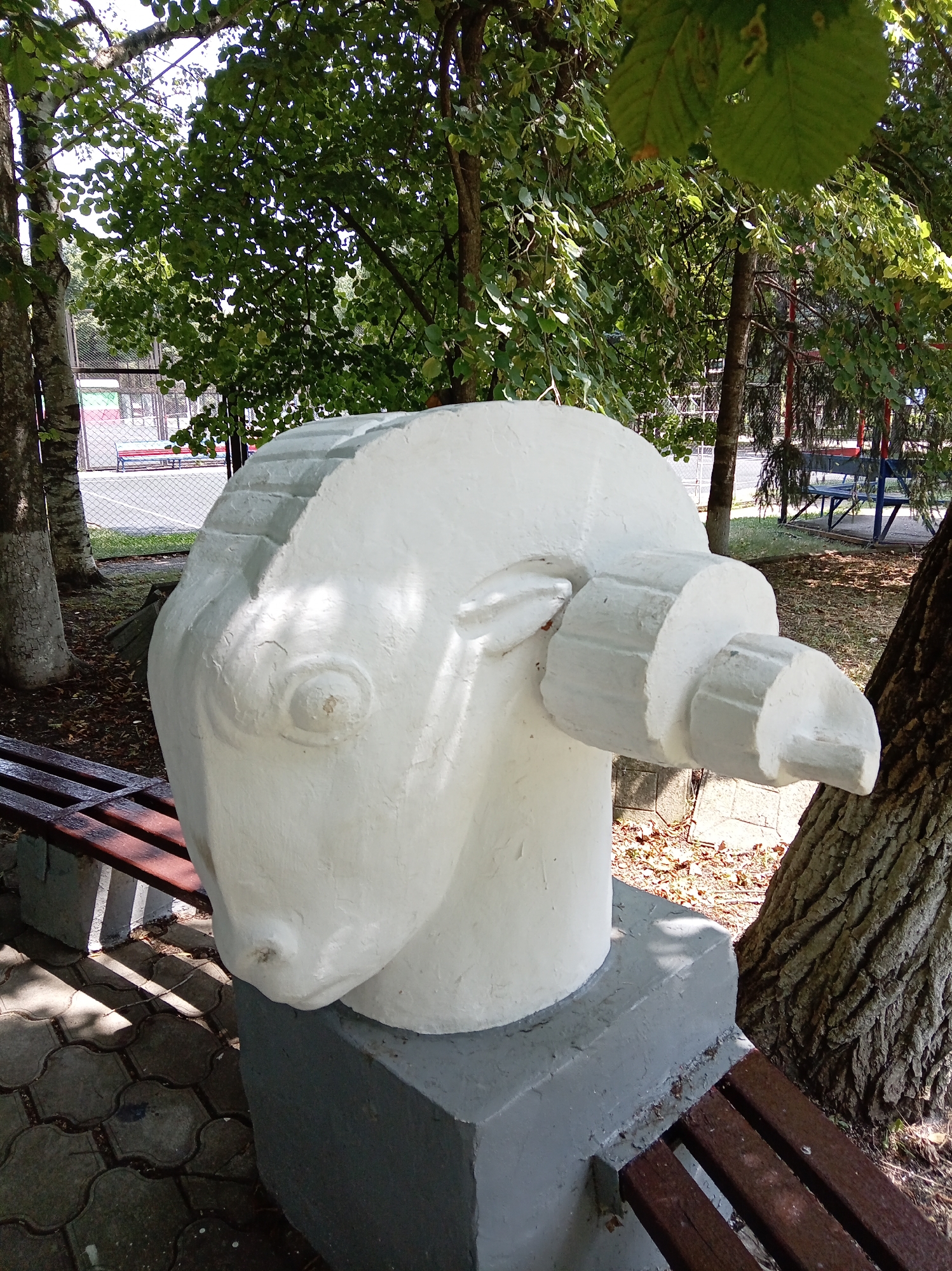 A white concrete sculpture resembling a stylized ram's head is situated outdoors in Yessentuki, Russia. The sculpture, mounted on a gray concrete pedestal, is positioned next to a wooden park bench with metal supports. Behind the sculpture, trees with lush green foliage create a shaded environment. In the background, a chain-link fence encloses a recreational area with colorful benches and playground equipment. The ground in front of the sculpture is paved with interlocking concrete tiles, and fallen leaves are scattered around the base. Sunlight filters through the trees, casting dappled shadows on the ground and the sculpture.