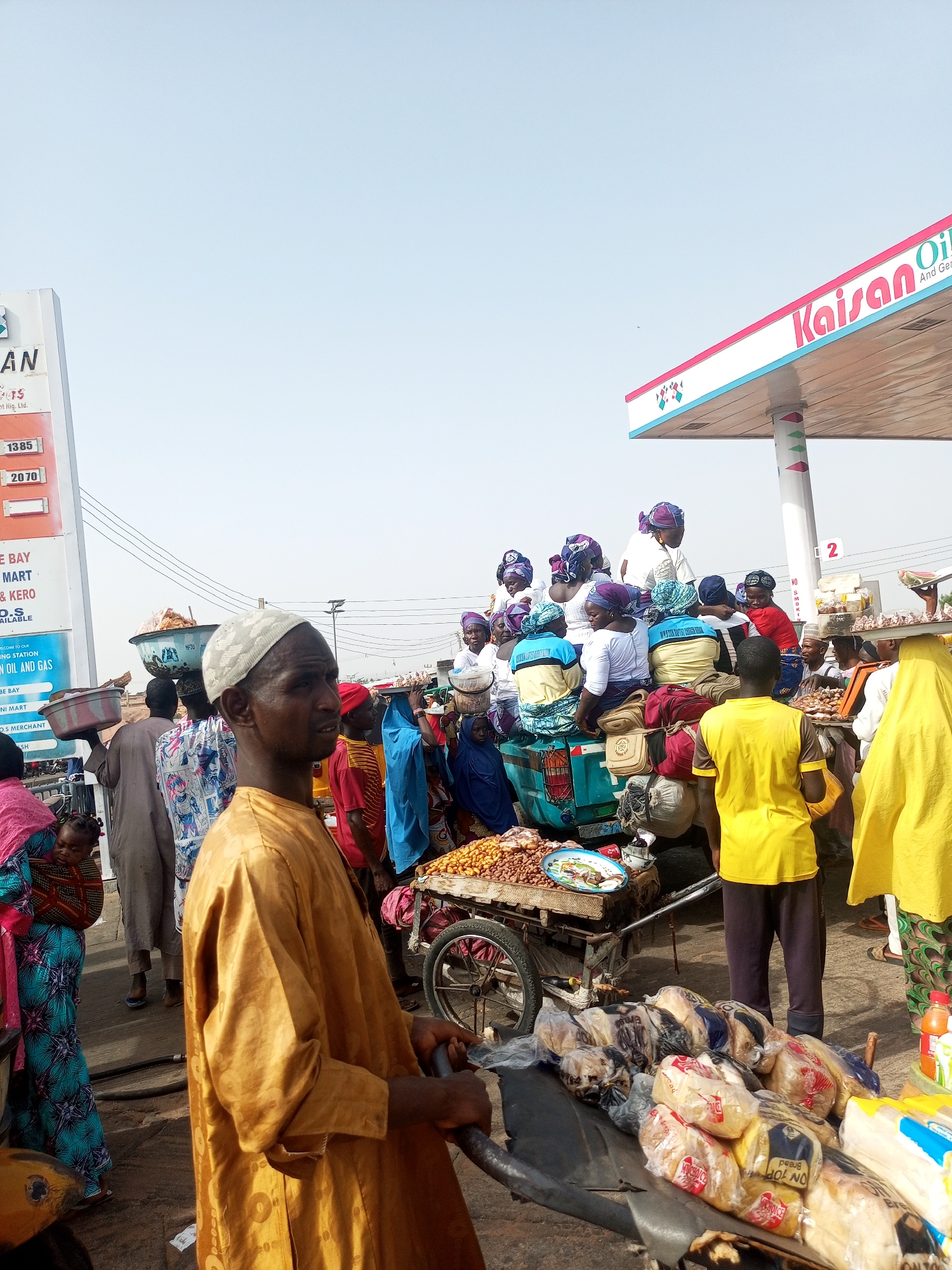 The image shows a bustling street scene in Gombe, Nigeria, near a Kaisan Oil and Gas station. A group of women wearing headscarves and colorful clothing are gathered in the back of a blue pickup truck. On the street, people are walking and selling goods, including bread and peanuts. A man wearing a yellow robe and a white cap is pushing a cart with bread on it. Other individuals are carrying items on their heads. The image captures a typical day in a busy commercial area of the city.