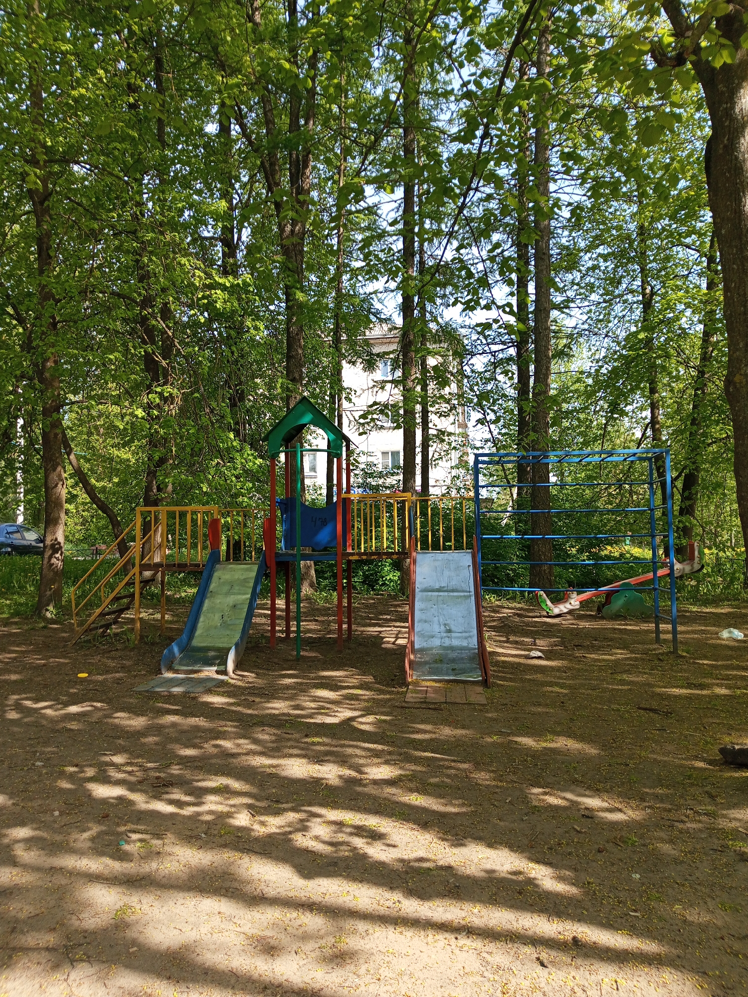 The image shows a playground located in Kommunar, Russia. The playground is situated in a wooded area, with tall trees surrounding the play equipment. The equipment includes two slides, a climbing structure with monkey bars, and a seesaw. The equipment is made of metal and plastic, with a variety of colors. The ground is dirt covered with fallen leaves and twigs, with patches of sunlight filtering through the trees. There is a building visible in the background, between the trees. A car is visible near the left edge of the frame. The playground appears to be empty.