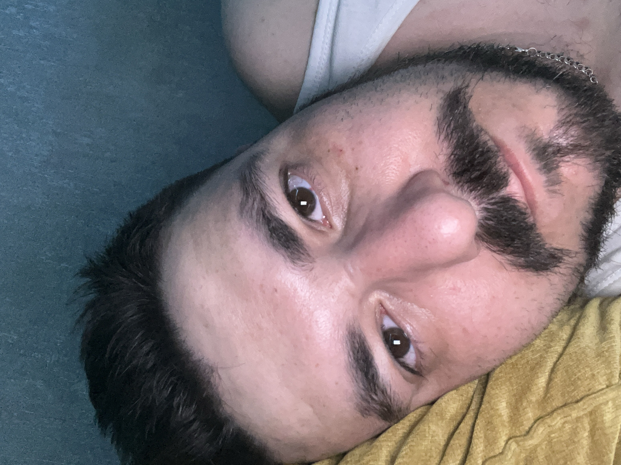 A close-up, angled selfie of a man. The man has brown eyes, dark hair, and a mustache/beard. He is wearing a white tank top and a silver chain necklace. He is lying on a yellow fabric with a blue surface behind him. There is no evidence that this photo was taken in Chaikovsky, Russia.
