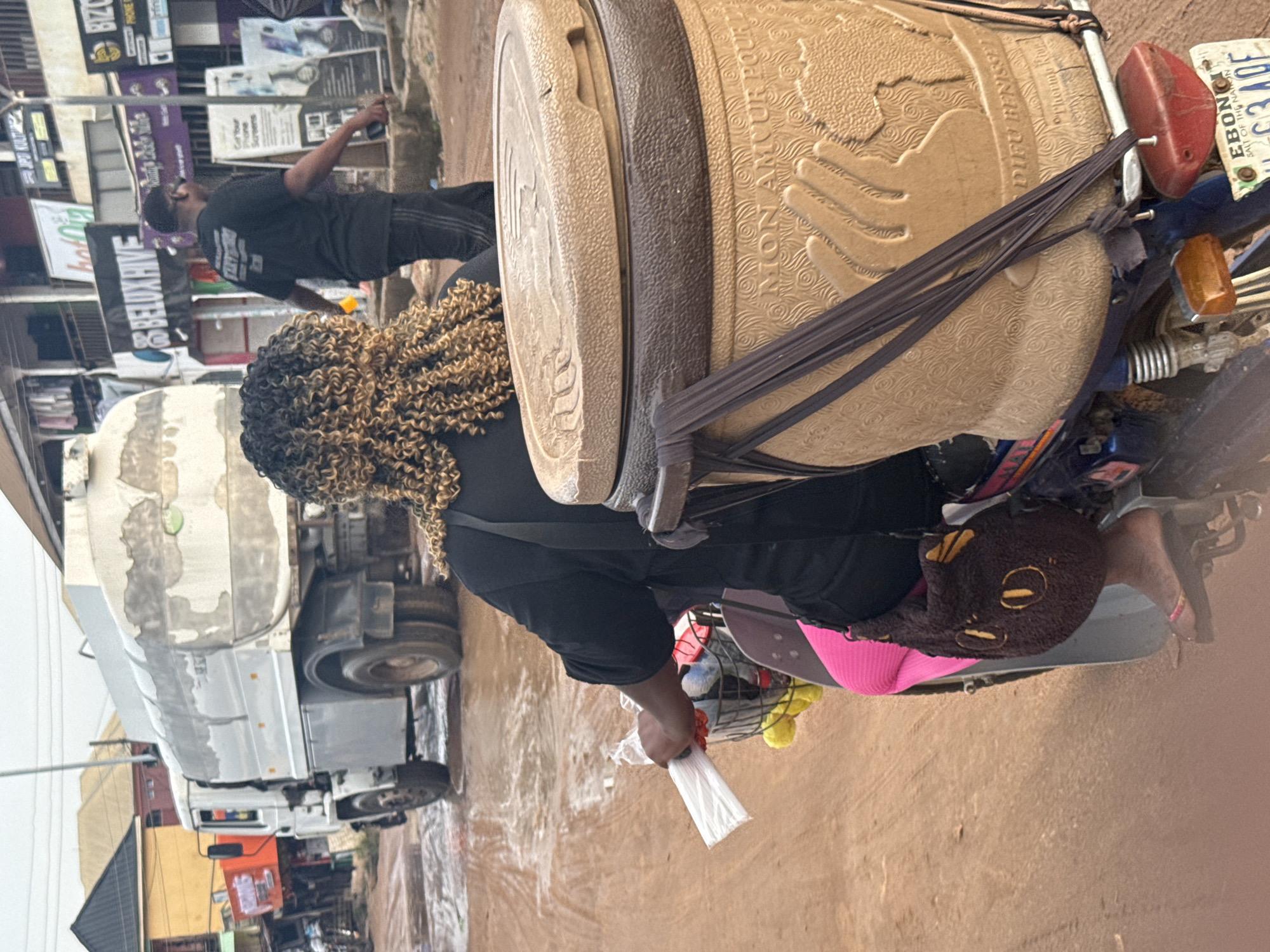 The scene depicts a woman with curly blonde hair riding a motorcycle in Ihiagwa, Nigeria. She is wearing a black shirt and a pink garment, carrying a large beige water container labeled "NON AMOUR POUR" and "DIEU BENIS". The container is secured to the bike with dark straps. The woman holds a plastic bag in her hand. The motorcycle is laden with other items, including what appears to be a stuffed animal.
In the background, a man in a black shirt is walking past storefronts with advertisements. A large white truck is also visible. Signage such as "BELUX HIVE", "BIZC", and "EBONGI SALT OF THE NATION" are evident. The ground is bare earth.