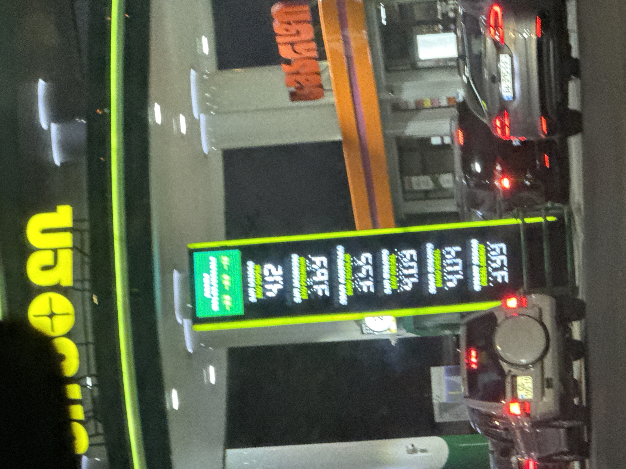 The image shows a gas station at night in Tbilisi, Georgia. A brightly lit vertical sign displays fuel prices, with numbers such as 4.12, 3.69, 3.55, and 4.09 visible. The gas station structure is illuminated with neon green lighting. Several cars are present, including an SUV with a spare tire mounted on the back, and a sedan. The sky is dark, indicating nighttime. The overall scene depicts a typical view of a gas station operating after dark.