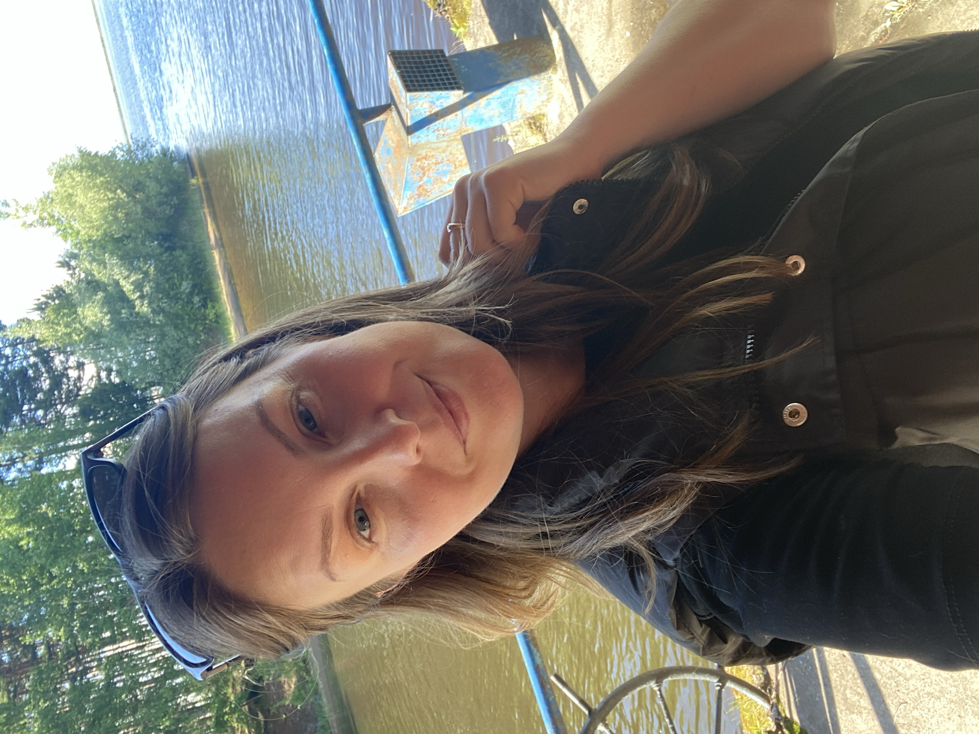 A woman is in Chaikovsky, Russia. She is on a boat or pier overlooking a body of water reflecting the sky and surrounding green trees. The woman wears a black jacket and sunglasses. She is fair-skinned with shoulder-length hair. She is smiling slightly at the camera. She wears a ring on her left ring finger. A metal grate is visible in the background. There is a blue railing.