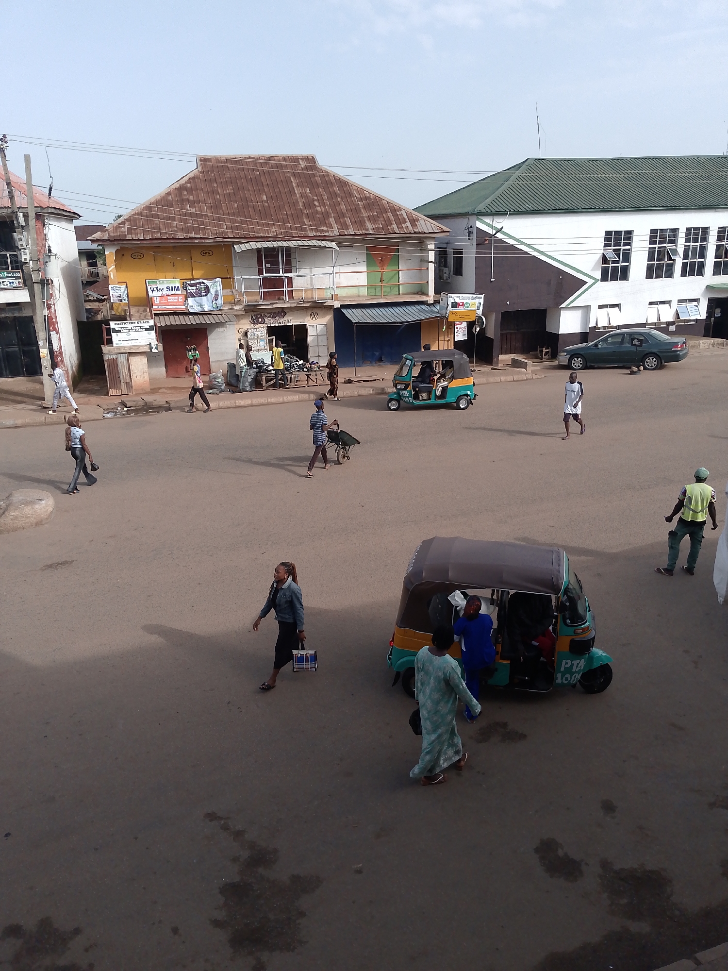 In Bukuru, Nigeria, a street scene unfolds with a two-story building featuring a brown, corrugated metal roof and yellow and white facade on the left, adjacent to another building with a similar roof and a white facade. To the right, a building with a green, corrugated metal roof and white and brown exterior walls stands prominently, with a dark green sedan parked in front of it. The street is paved with asphalt and shows signs of wear, including some dark wet patches.
Several people are visible on the street. Three tricycles, or "keke napeps," are present: one teal and yellow in the middle of the street, another teal and yellow with "PTA 100" printed on its side in the lower right portion of the image, and a third teal and yellow tricycle near the left side of the image.
A woman in a blue denim jacket and black skirt walks towards the lower right corner carrying a blue and white bag. Another person, dressed in a light blue and white patterned garment, walks towards the left, appearing to step off the curb. A man in a yellow safety vest and green pants stands with his arms extended, facing away from the camera. A person in a white t-shirt and dark shorts runs across the street in the mid-ground, and another individual in a striped blue and white shirt pushes a wheelbarrow from right to left in the mid-ground. Several other figures are scattered throughout the scene, engaged in various activities such as walking and interacting. The sky above is a pale blue with some overcast.