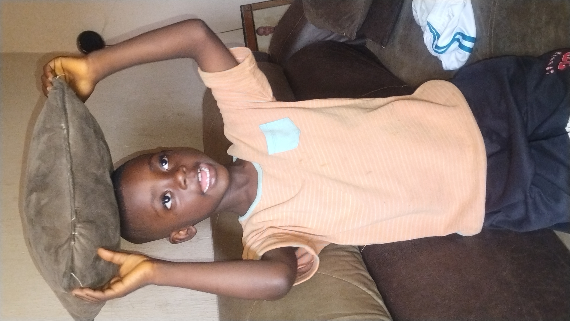 A young boy is balancing a brown pillow on his head. He is wearing a peach-colored striped t-shirt with a light blue pocket and dark blue shorts. He is standing indoors, likely in a living room. The background shows a brown couch and a framed picture of a man. The lighting suggests it is daytime. The overall scene appears to be a candid moment.