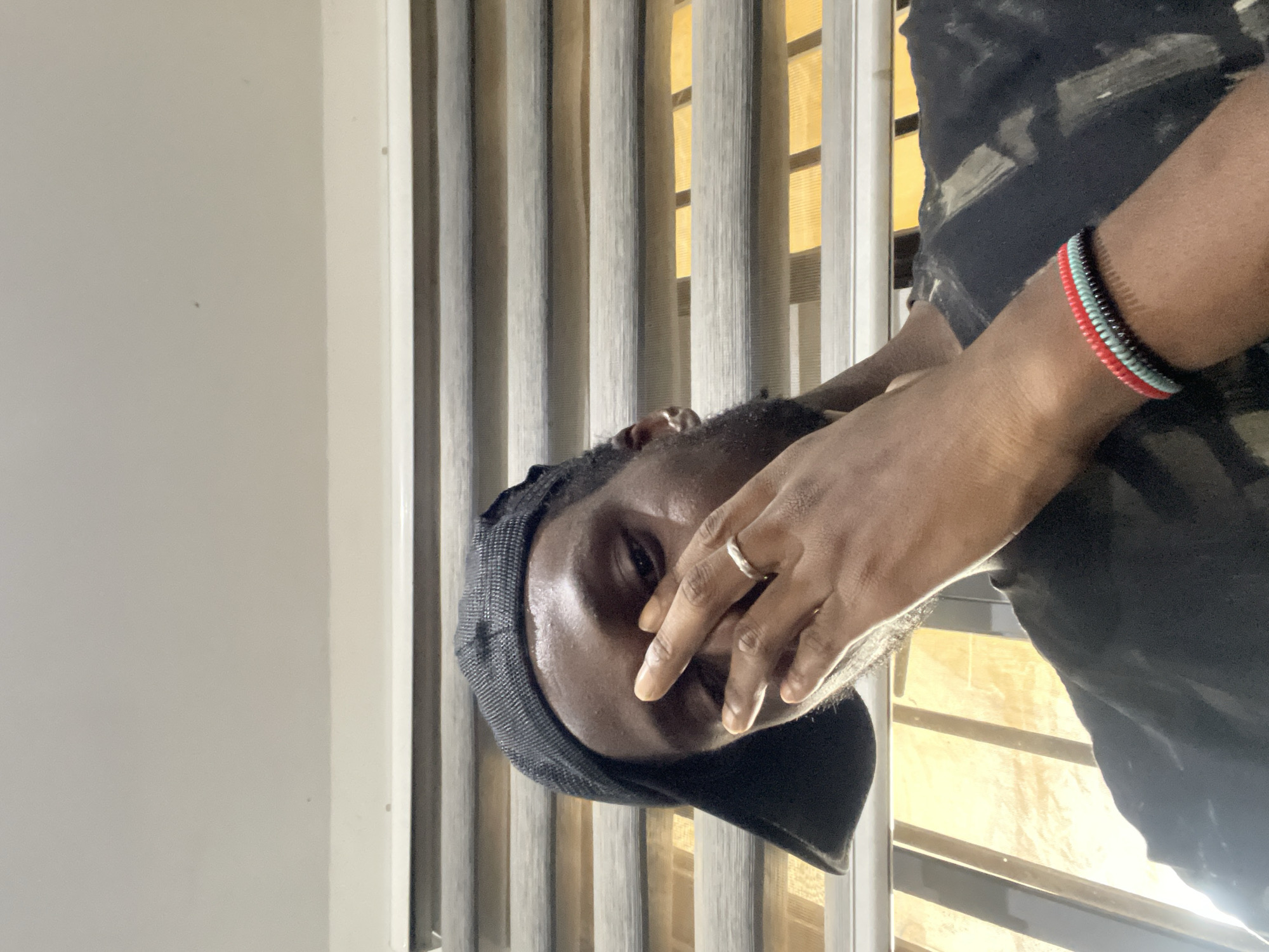 The image is a close-up, selfie-style shot of a Black male wearing a black baseball cap and a black t-shirt with a camouflage pattern. He is indoors, possibly in a residential setting in Abuja, Nigeria, as suggested by the distinctive horizontal blinds with thin, light-colored slats and thicker, darker slats that are partially open, revealing a yellowish light source behind them. The man is looking towards the camera, and his right hand is covering the lower half of his face, with his index finger and thumb touching his nose. He is wearing a silver band ring on his left ring finger and a woven bracelet with red, light blue, and black bands on his left wrist. The lighting is dim, with a bright light source visible on the left side of the frame, possibly indicating a window or lamp. The background consists of the horizontal blinds and a lightly colored wall above them.