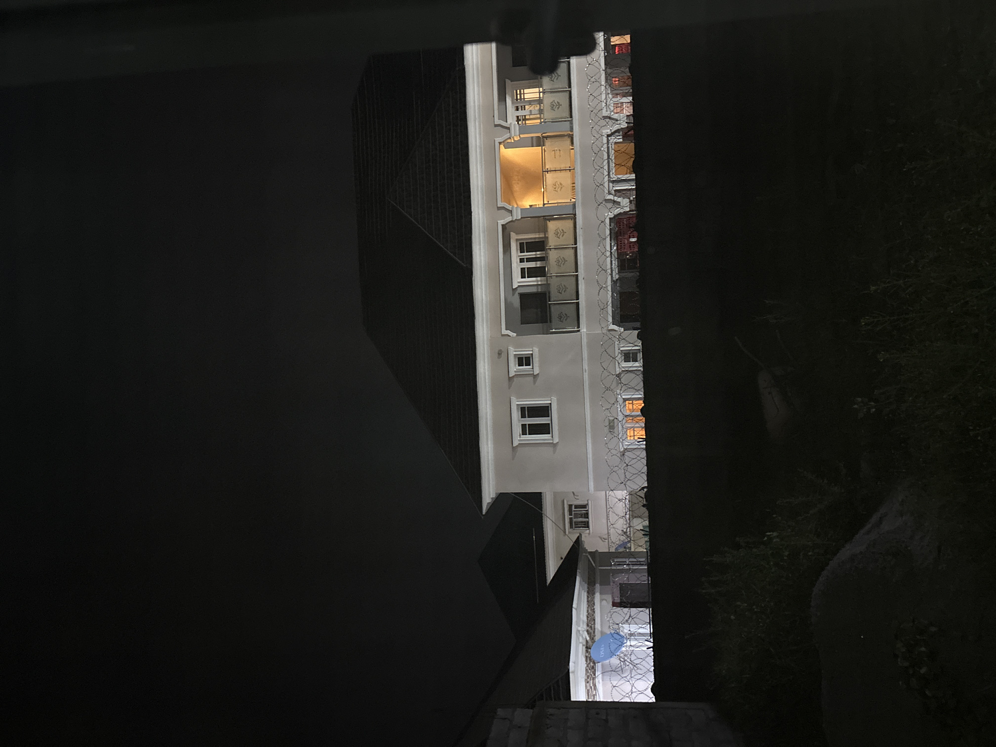 The image shows a modern residential building at night in Angwan Sayawa, Nigeria. The building is multi-story with a dark-colored roof and light-colored exterior walls. There are several windows visible, some of which are illuminated from within. A balcony with a decorative railing is visible on the upper floor, with warm interior lighting. A fence, possibly made of barbed wire, runs across the foreground, partially obscuring the view of the building. To the left of the main building, another structure with a similar roofline is visible. The sky is dark, indicating it is nighttime. The foreground is obscured by darkness, with some vegetation and a rocky area visible.