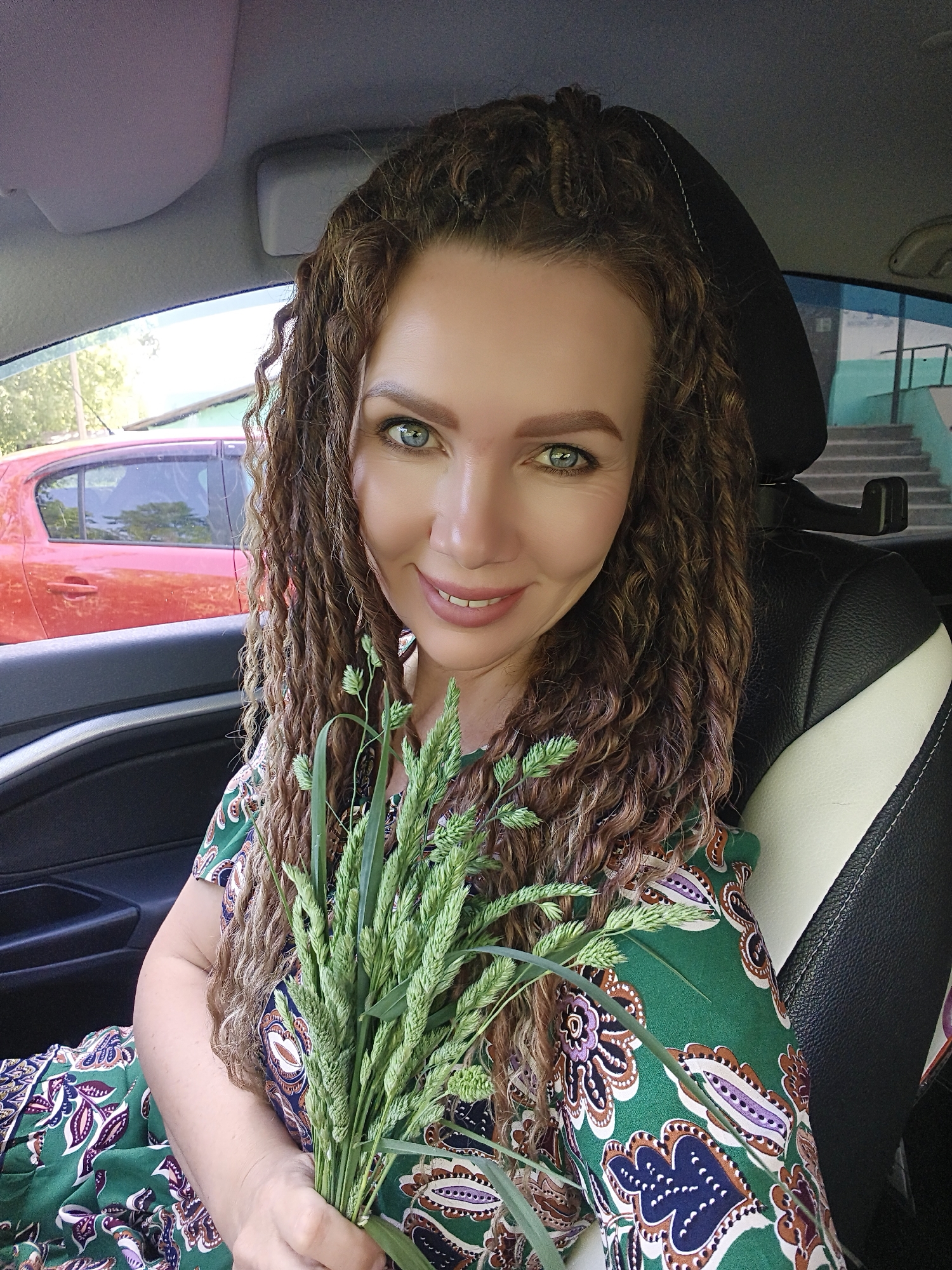 A woman with curly brown hair and green eyes is holding a bunch of tall green grass in her hands. She is sitting in the driver's seat of a car with black and white leather seats. The interior of the car is visible with a grey roof and dark door panels. Outside the car, a red car is parked and a building with a turquoise painted wall and a set of stairs leading up to a doorway is seen. The exact location is Unknown, Unknown. The woman is smiling at the camera.