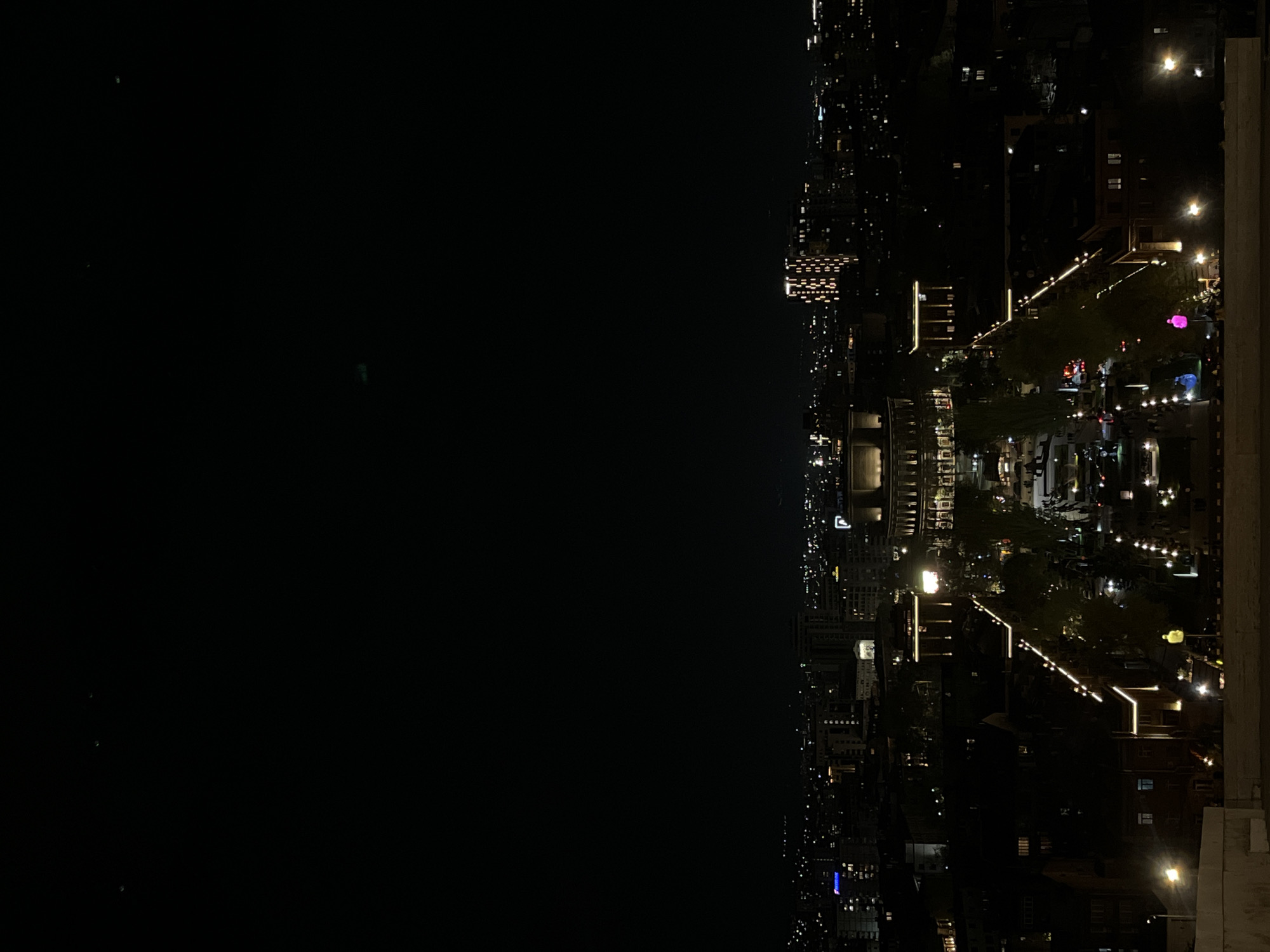 The image displays a sprawling cityscape at night, viewed from a high vantage point. The scene is dominated by artificial lights against a dark sky.
In the lower portion of the frame, numerous buildings are visible, illuminated by their interior lights and streetlights. A prominent feature is a complex system of what appear to be multi-level terraces or steps, possibly part of a park or a large public area, brightly outlined with decorative string lights and architectural lighting along railings and pathways. These illuminated structures zigzag across the mid-ground, leading to what might be a central plaza or open space with more lights. Patches of greenery, likely trees, are interspersed among the buildings and pathways, some also adorned with lights. A few distinct colored lights, such as a pink glowing object and a blue light, add splashes of color to the urban landscape.
The upper portion of the image is largely dark, suggesting a vast expanse of unlit land, water, or the night sky, with only a few faint, distant lights barely discernible. The overall impression is one of a vibrant, illuminated city after dark. The time of day is clearly night, and the sky appears clear. There are no clearly identifiable individuals or vehicles in detail. The image provides no visible text. Given the location context, this is a night view of Yerevan, Armenia.