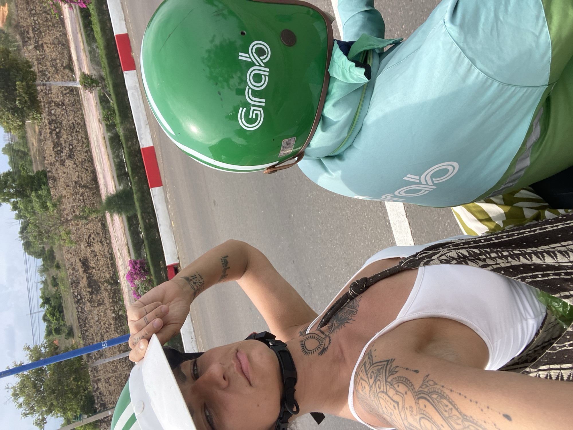 In this medium shot taken from behind and to the side of two individuals on a scooter, the focus is on the two people and their attire as they travel down a paved road in Phu Thuy Ward, Vietnam. The person in the foreground, on the left, is a woman wearing a white helmet and a white tank top, revealing extensive tattoos on her arm, chest, and neck. Her left hand is raised, adjusting the brim of her helmet. To her right, the second person, likely a man given the attire, wears a green Grab helmet and a light blue jacket with the Grab logo visible on the back. The jacket appears to be part of a uniform, possibly for a delivery driver. The background shows a stone wall with some greenery and trees, characteristic of a roadside landscape in Vietnam. The sky is overcast, suggesting a warm, humid climate. The road is asphalt and appears to be in good condition, with a red and white painted curb visible on the right side of the image.