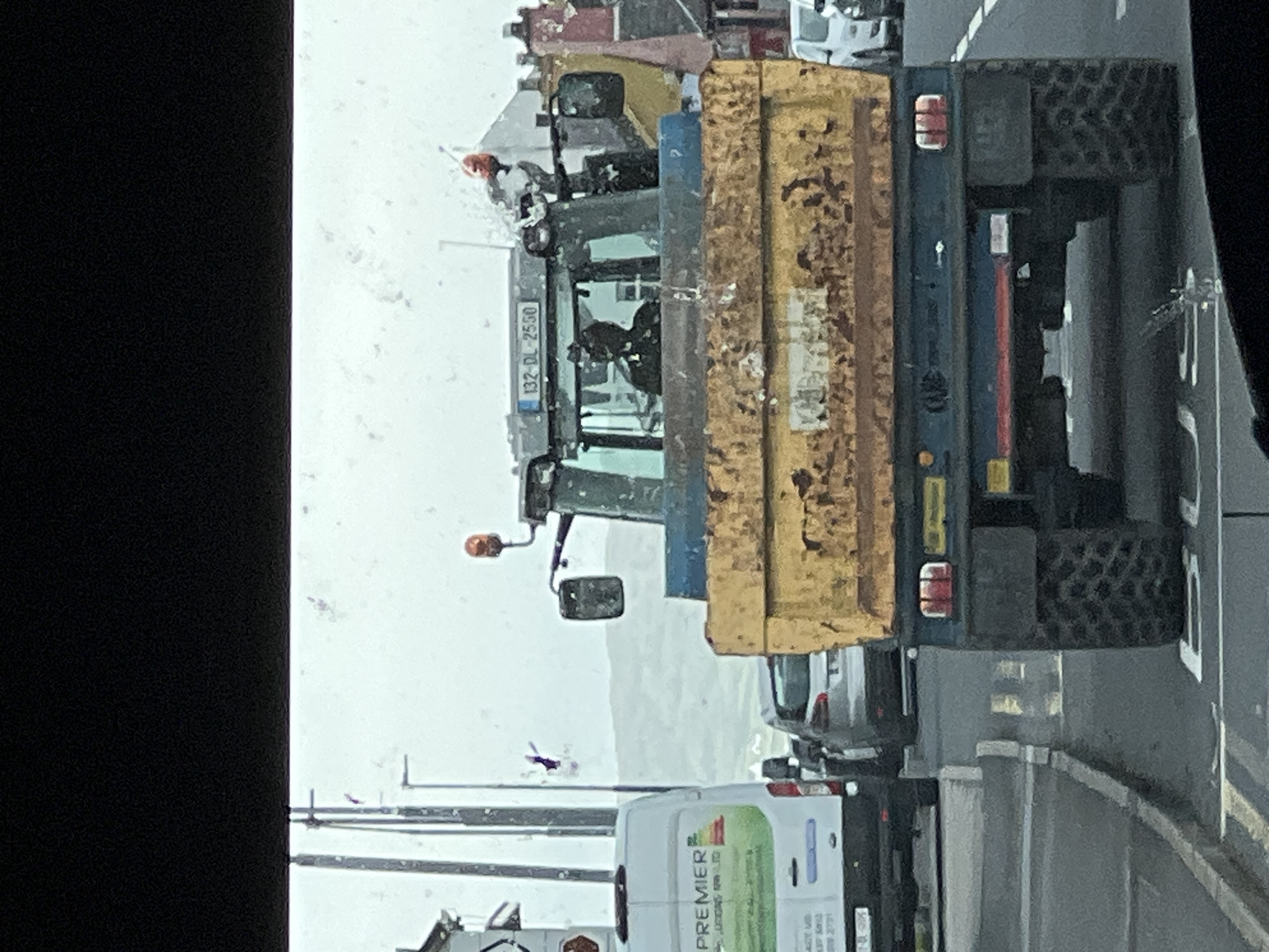 The image shows a close-up, slightly angled view of a large yellow and blue tractor, likely a snow plow given the weather. The tractor is on a road, which appears wet and possibly icy. Snowflakes are visible in the air, indicating it is currently snowing or has recently snowed. The scene suggests a cold, overcast day.
The tractor has a large yellow hopper at the back, likely for carrying salt or snow. Its front features a snowplow attachment. A license plate on the front of the tractor reads "132-DL-2550". The driver's cabin is visible, with a person seated inside. Other vehicles are partially visible behind the tractor, including a white van with "PREMIER" and "LANDSCAPES" written on its side, suggesting a possible service vehicle. The road has white lines indicating lanes.
The overall setting appears to be a rural or semi-urban area, with the presence of the tractor suggesting road maintenance in response to adverse weather conditions. The limited visibility due to the snow and overcast sky contribute to a somber, functional atmosphere. The image seems to be taken from inside another vehicle, as indicated by the black border on the left and the perspective. The location is identified as Falcarragh, Ireland.
