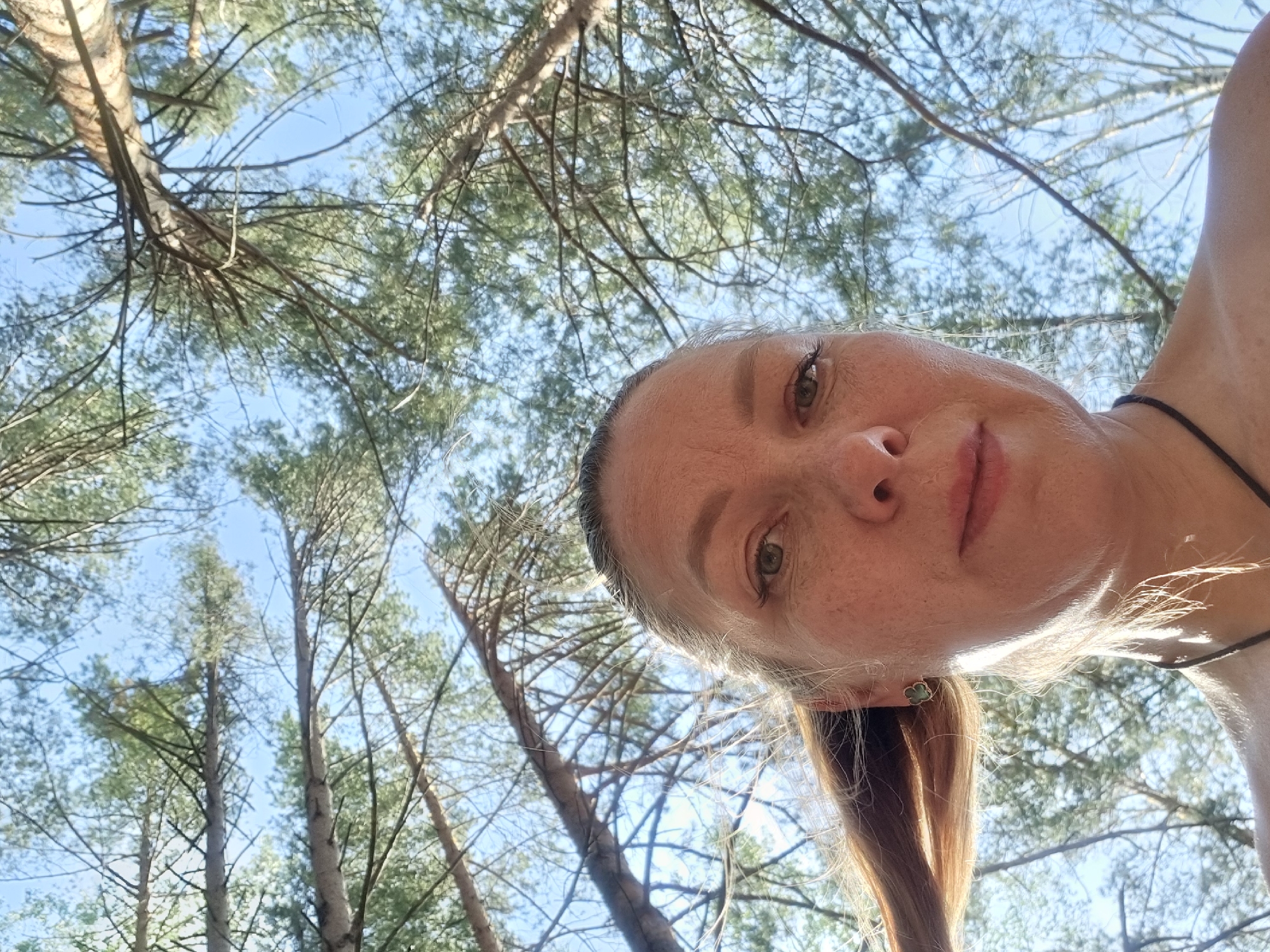 The image is a low-angle selfie of a fair-skinned woman with reddish-blonde hair. Her head is tilted towards the right, and her face is the primary subject. She has freckles and is wearing a small, green earring. A thin black necklace is visible around her neck. The background consists of tall trees reaching into a bright, blue sky. The trees appear to be in a forest or wooded area. Due to the lack of distinguishing landmarks, providing a precise location within the city of Chaykovsky, Russia is not possible.