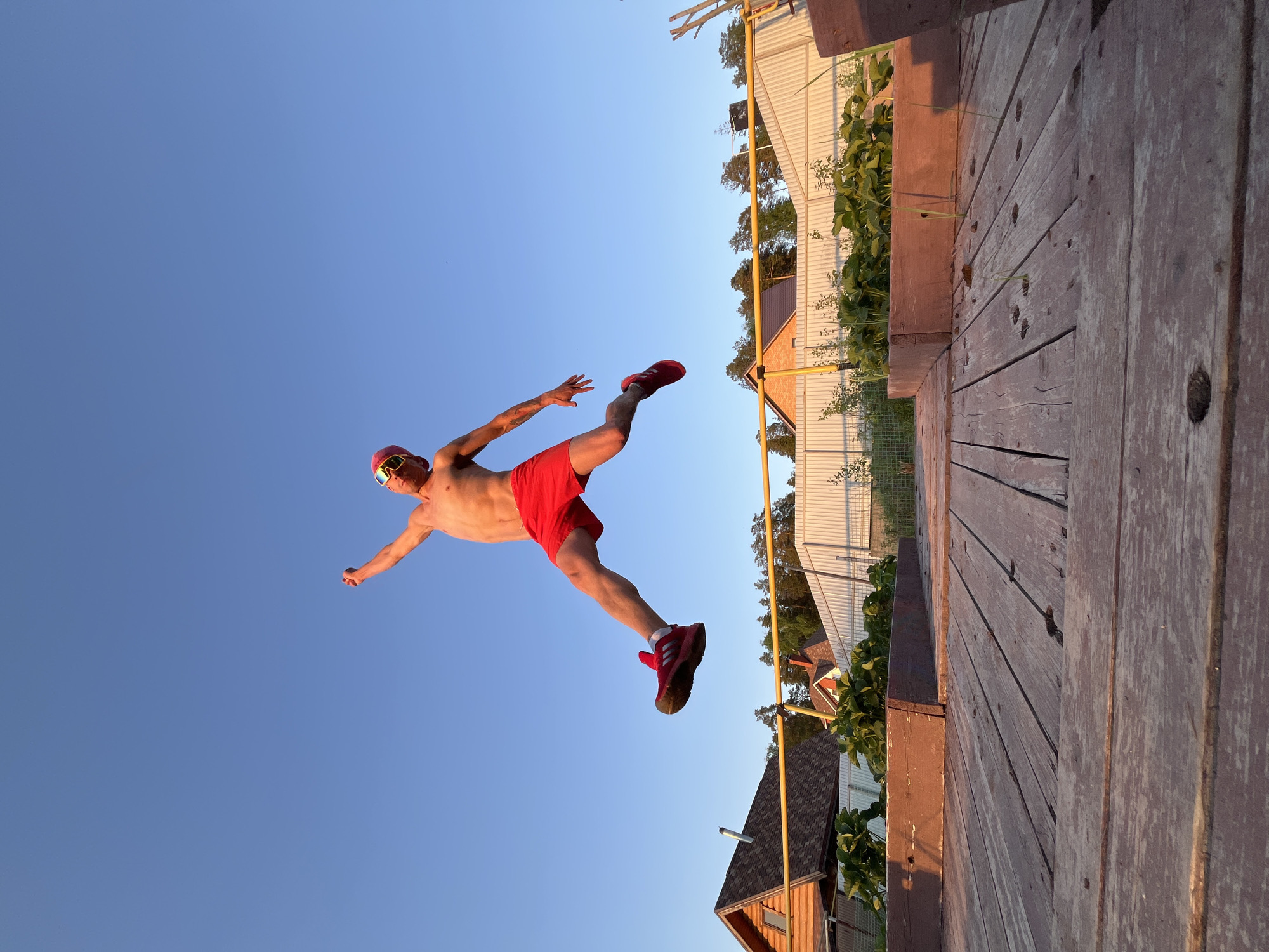 A shirtless man wearing red shorts, red shoes, and sunglasses is airborne against a clear blue sky. He is positioned at an angle, appearing to leap toward a wooden platform or deck. To the right of the deck, glimpses of houses with pitched roofs and greenery are visible. The photo was taken in Chaikovsky, Russia.