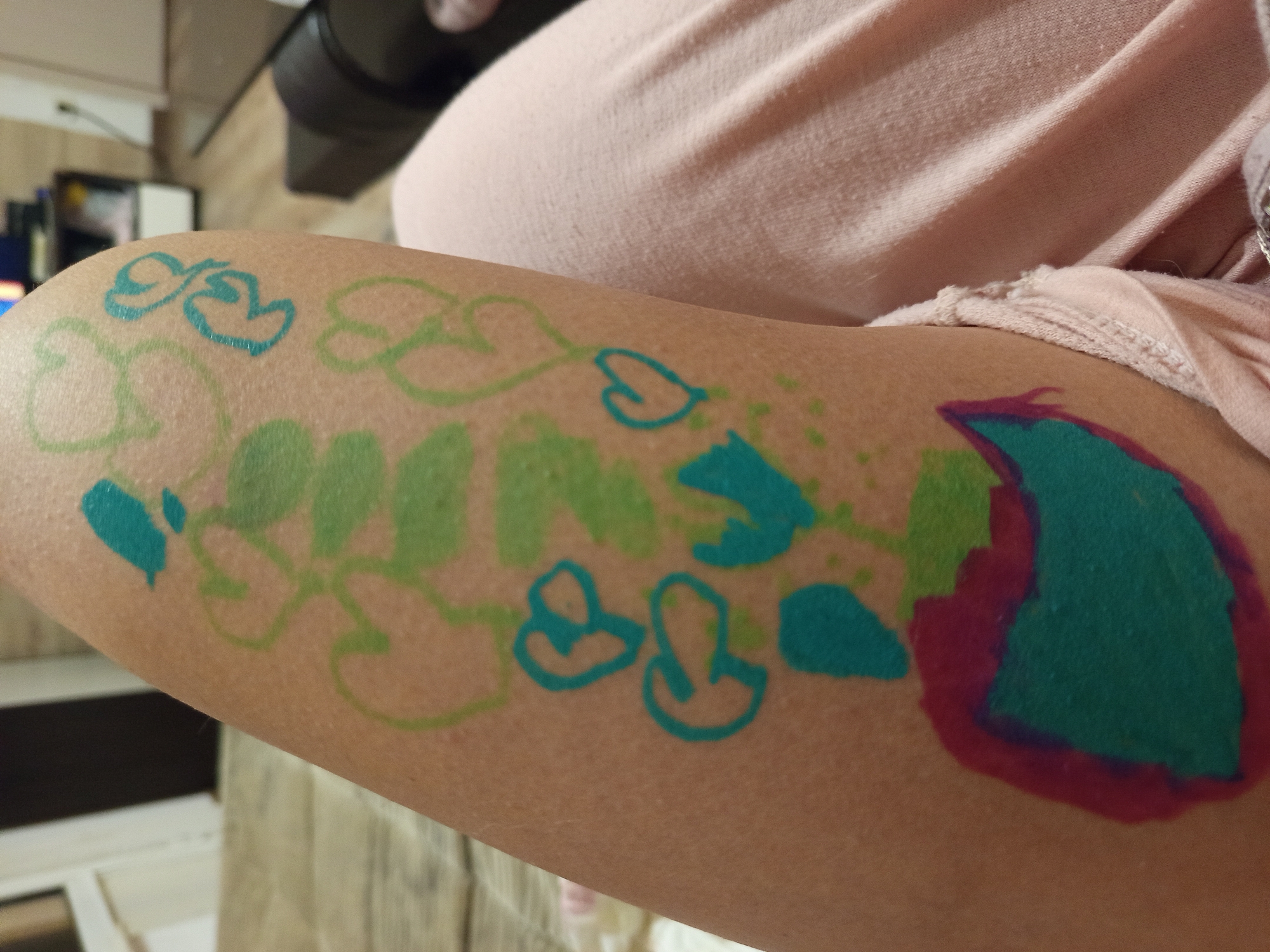 An arm is visible, adorned with drawings in blue, green, and purple markers. The drawings appear to depict a vine with leaves, hearts, and a larger, amorphous shape outlined in purple and filled with blue. The arm is positioned against a background that includes light-colored clothing, a countertop, and blurry objects in the distance. The location is not discernible from this image.
