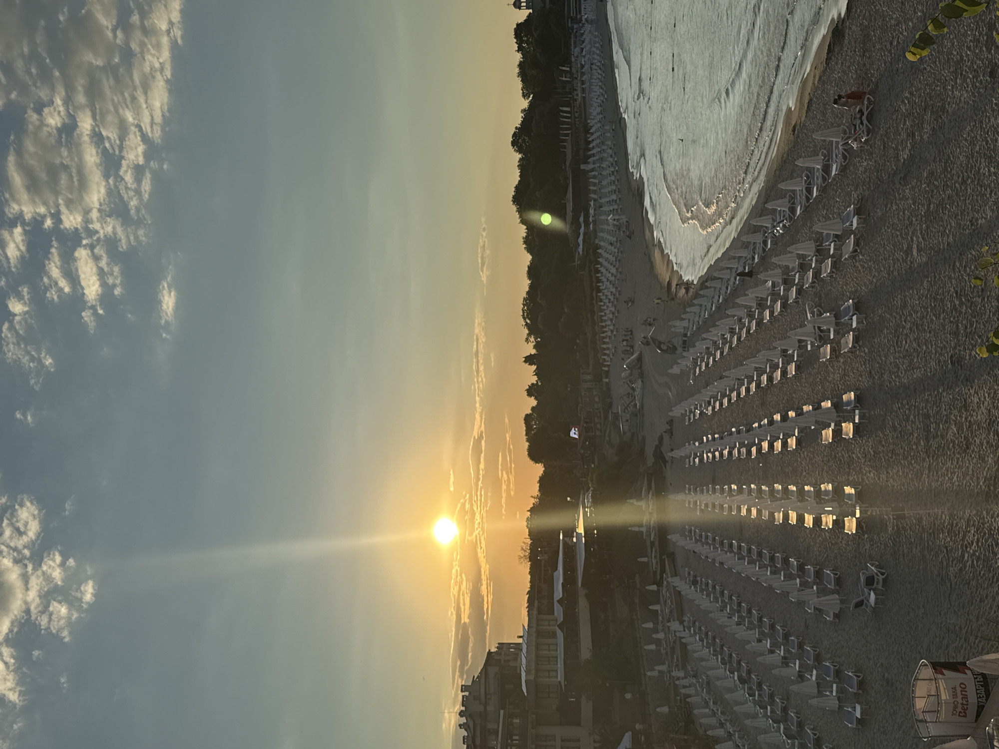The image depicts a sunset view of the beach in Sozopol, Bulgaria, specifically overlooking the coastline from an elevated position. The sky is a gradient of blue and orange, with scattered clouds illuminated by the setting sun, which is a bright, circular orb positioned in the upper left quadrant, casting a strong light ray downwards.
The beach is vast and appears to be mostly empty, with numerous rows of neatly arranged beach chairs and umbrellas, most of which are white. The sand is a light brown color. The sea is visible on the right, with gentle waves rolling onto the shore, reflecting the warm light of the sunset.
Along the left side of the image, several buildings are visible, appearing to be hotels or resorts, with architectural elements suggesting a Mediterranean or Eastern European style. These buildings are partially silhouetted against the sky and the landscape. Behind the beach, a dense line of green trees forms a natural backdrop, with some taller structures, possibly including a church steeple, visible amidst the foliage.
A few people are scattered on the beach and near the water's edge. On the far right, two individuals are near beach chairs, one appearing to be seated and the other standing. Further down the beach, small groups of people are seen walking or relaxing. In the foreground, a white cylindrical object with some text on it, possibly an advertisement or a trash receptacle, is visible on the left. The overall scene conveys a sense of tranquility and the end of a day at the seaside.