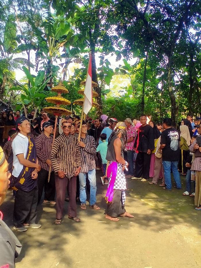 The image shows a group of people gathered outdoors in Masaran, Indonesia (latitude: -7.471, longitude: 110.957). The group consists of mostly men, all wearing traditional Indonesian clothing. There are several women present, also wearing traditional clothing. Some are holding a large Indonesian flag, while others hold parasols adorned with woven straw. The environment is lush and green, with mature trees creating a shaded canopy over the gathering. The ground is paved and dusty, with areas of grass and foliage. The group appears to be attending a traditional ceremony, possibly a wedding or a cultural celebration. The people are standing together in a group, some appear to be talking or interacting with each other, while others are standing still.