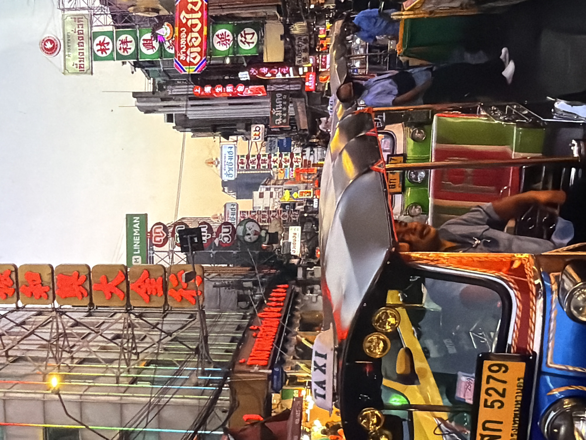 The image captures a vibrant street scene at dusk or evening, characterized by numerous illuminated signs in Thai and Chinese scripts. In the foreground, a blue and yellow tuk-tuk is prominently featured, adorned with intricate gold and red detailing. Its driver, a man with a relaxed expression and slight smile, is visible behind the wheel. The tuk-tuk's license plate displays "สก 5279 กรุงเทพมหานคร" (SK 5279 Bangkok) and "2569", along with a small "TAXI" sign. Further down the street, another green and red tuk-tuk is visible, with a person lying down in the passenger area, possibly resting.
The narrow street is lined with multi-story buildings, densely covered with a variety of brightly lit commercial signs. Visible Thai text includes "LINEMAN", "เลี้ยงเฮงตั้งที่", "ฮั่วเซ็งเฮง", "ห้างทอง", "ฉ.มั่นคง", "ชิงเฮง", and "ต้องใช้". Chinese characters like "林祥興" (Lin Xiang Xing) and "藥行" (Pharmacy/Medicine Shop) are also prominent on several signs. Overhead, a tangle of electrical wires crisscrosses the sky. The scene depicts a bustling urban environment, with other people visible in the background, suggesting ongoing daily activities. The image is situated in Lovech, Bulgaria.