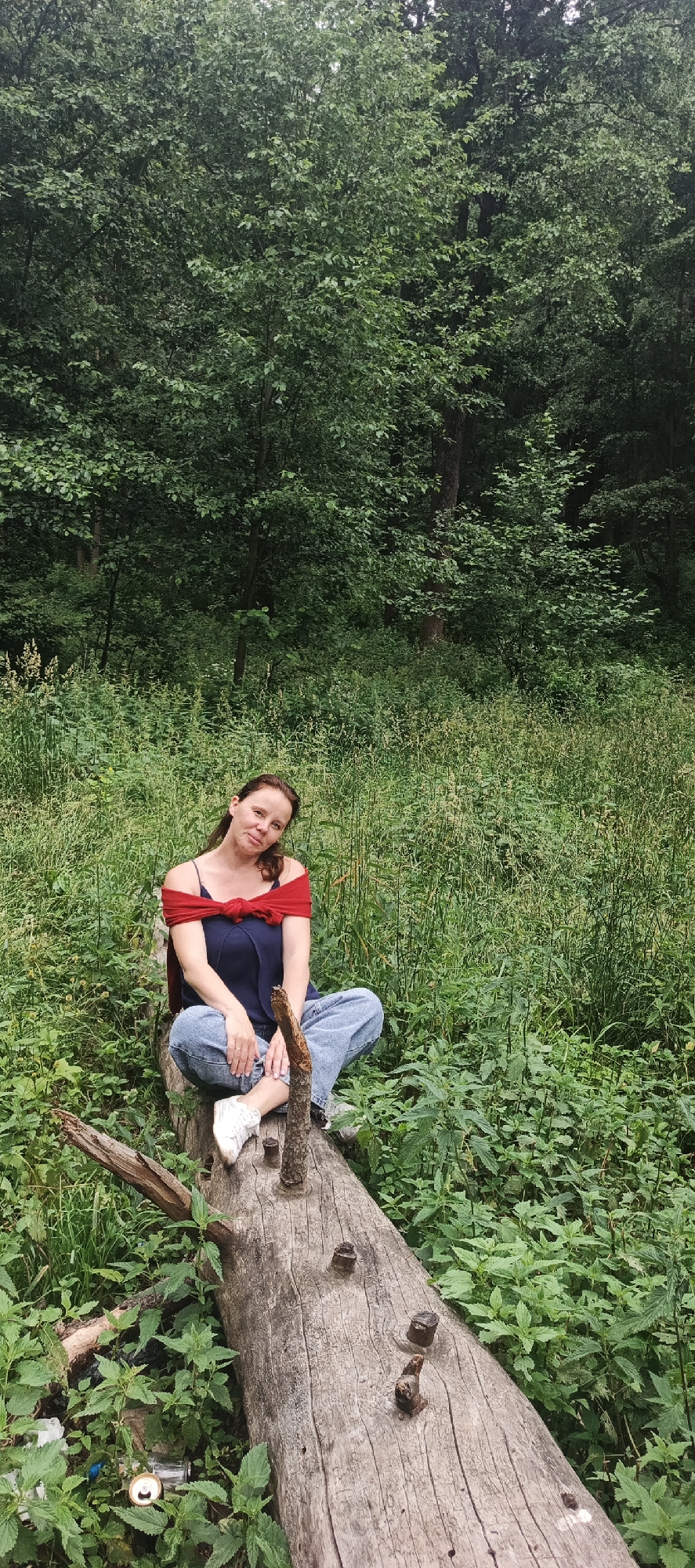 A medium shot shows a woman sitting cross-legged on a fallen tree trunk in a densely vegetated area. She is wearing a dark blue top with a red scarf tied around her shoulders, blue jeans, and white sneakers. Her hair is brown and pulled back. The tree trunk is rough and weathered, with several protruding branches. The background is filled with lush green trees and undergrowth, suggesting a natural, possibly wooded, outdoor setting. The lighting is soft, indicating an overcast day or diffused sunlight. The overall impression is one of a casual, serene moment in nature. The image does not contain any specific details that would allow for identification of the location within the city of Chaikovsky, Russia.