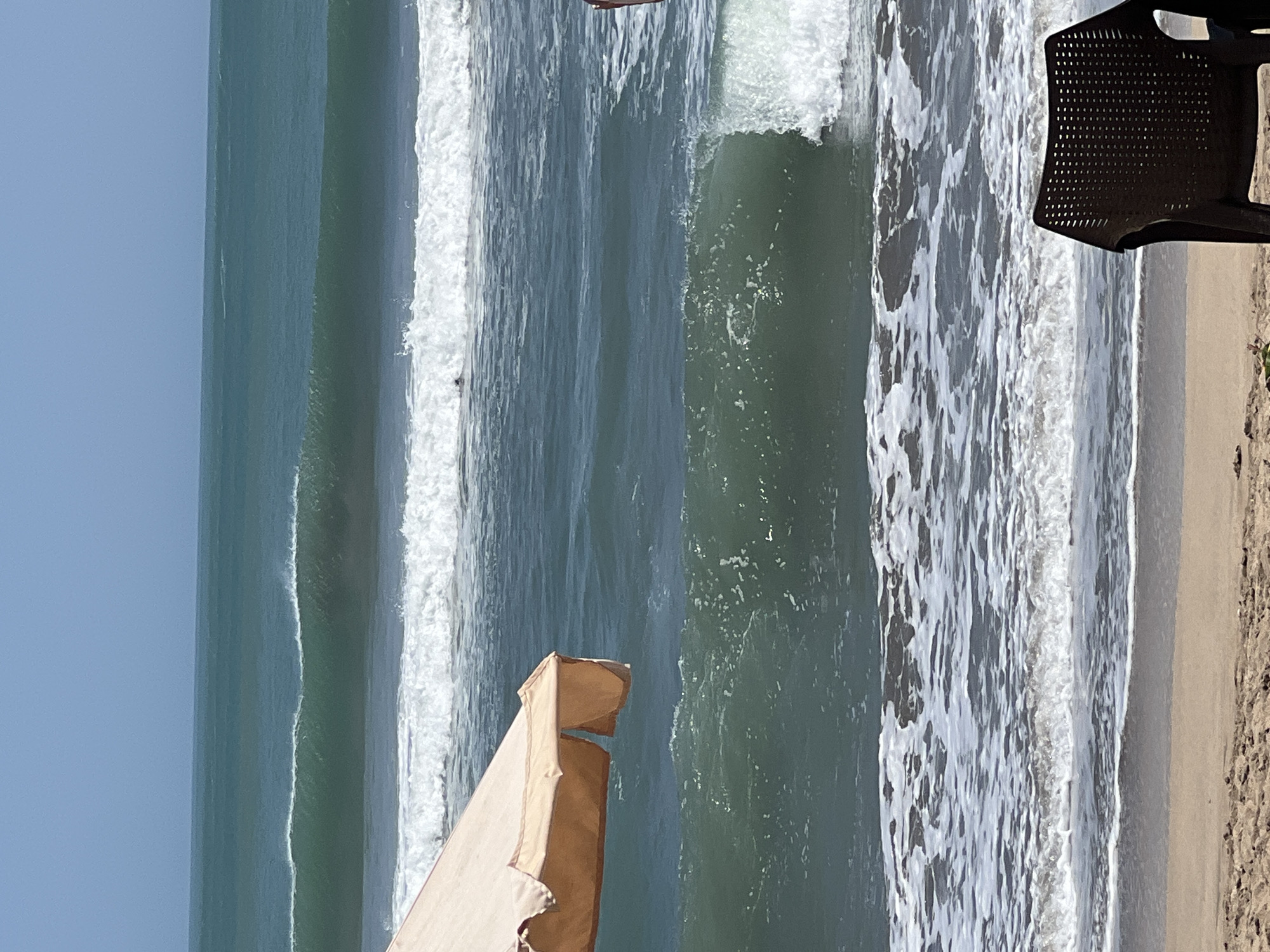 An ocean view at Kuta beach shows several sets of waves rolling in under a clear blue sky. In the foreground, a tan beach umbrella is partially visible on the left, and a black plastic chair faces the ocean on the right. The sand is light brown and textured. The water is a deep teal green, with distinct lines of white foam from the breaking waves. A solitary figure is visible swimming in the ocean between the second and third sets of waves from the shore.