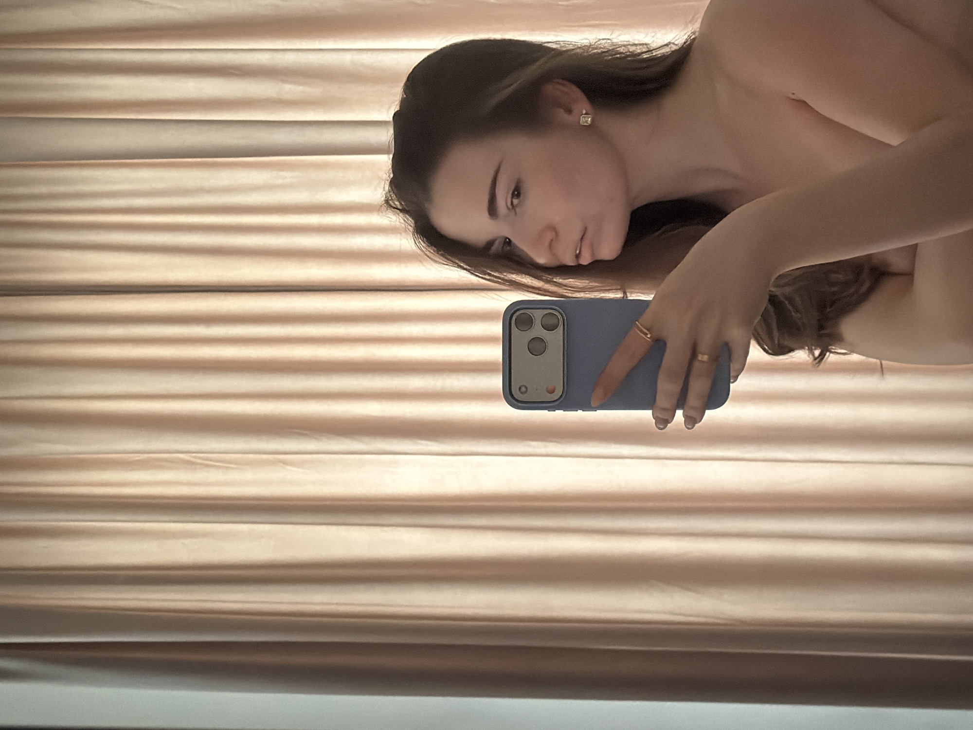 A woman is taking a selfie with a blue smartphone in front of a horizontally oriented beige curtain. The location is not identifiable as Moscow, Russia.