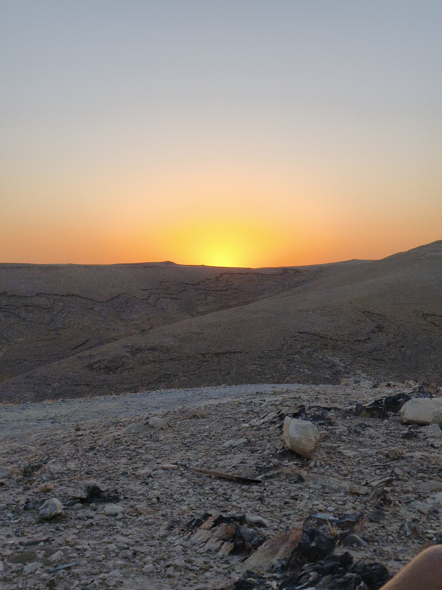 A wide-angle shot captures a desert landscape at dusk, with the sun setting on the horizon. The sky transitions from a pale blue at the top to a vibrant orange and yellow as it nears the setting sun, which is partially obscured by the distant hills. The terrain in the foreground is rocky and barren, with small stones and larger rocks scattered across the uneven ground. In the middle ground, rolling hills with a dusty, textured surface are visible, crisscrossed by faint lines that suggest erosion or dried water channels. The overall impression is one of a remote and arid environment, characteristic of the Keidar area in the Palestinian Territories. No people are visible in the frame.
