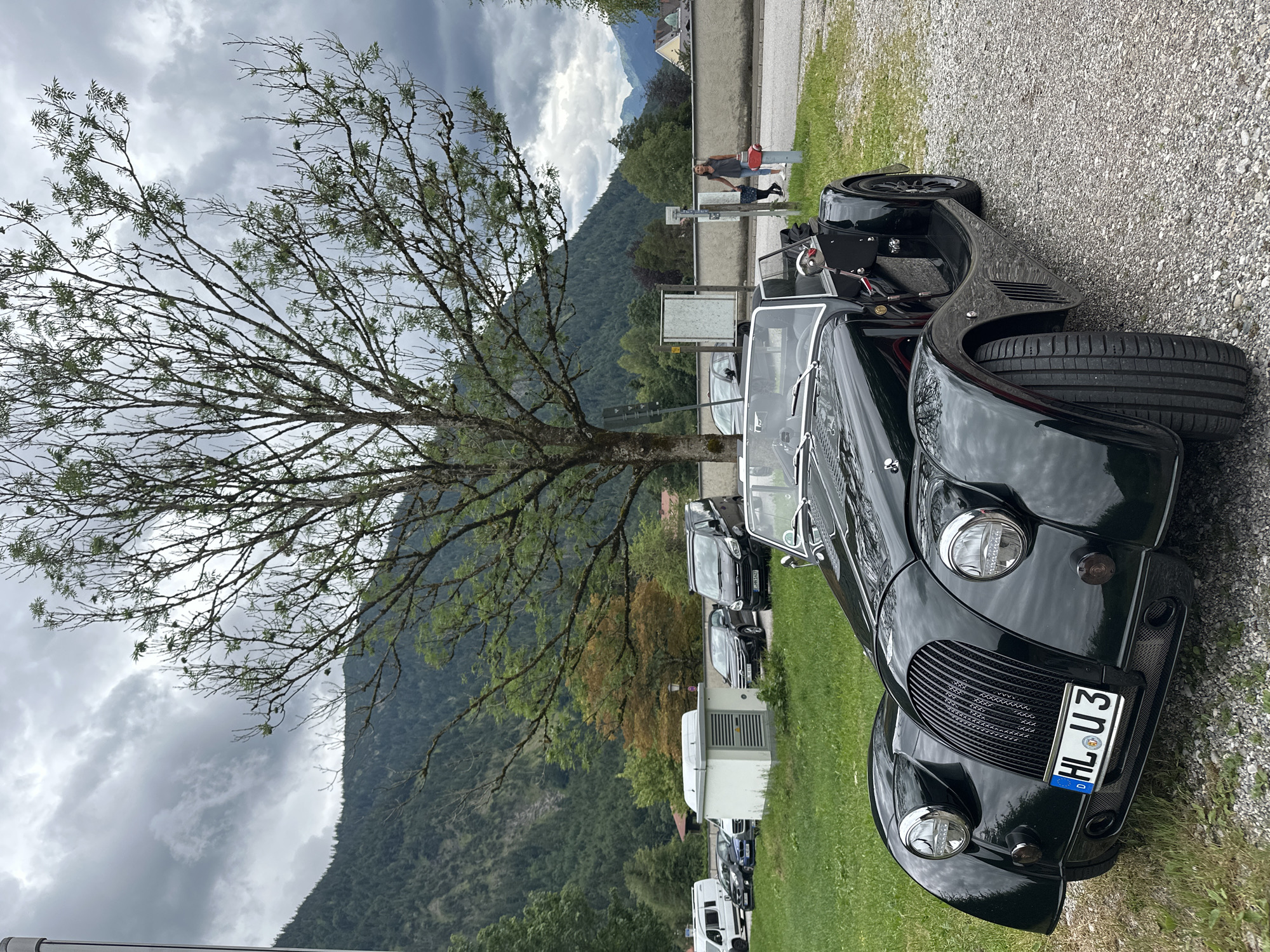 In Ettal, Germany, a dark green vintage convertible sports car is parked on a gravel surface in front of a grassy area. The car's front features a prominent grille, round headlights, and a license plate reading "HL U 3". In the background, a large tree with sparse branches stands against a cloudy sky. Beyond the tree, a mountainous landscape with lush green slopes and scattered buildings is visible. Several other vehicles, including a white van and several dark cars, are parked further back. A woman in a gray tank top and blue jeans is walking away from a red fire hydrant near a stone wall. The scene depicts an outdoor parking area in a scenic, mountainous region.