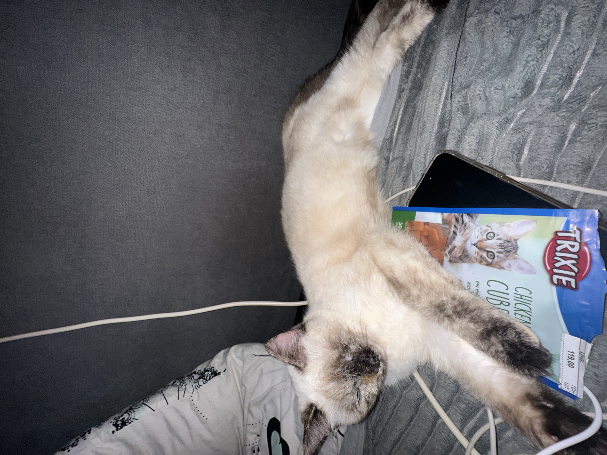 A Siamese cat is lying on a gray fuzzy blanket with its body stretched out. The cat is cream-colored with dark brown points on its ears, face, and paws. It is resting on top of a package of "TRIXIE CHICKEN CUBES" and a black smartphone. A white cord runs across the blanket and the cat's body. In the background, a dark gray upholstered surface is visible, as well as a white pillow with a black and white patterned design. The scene appears to be indoors, possibly in a bedroom or living room, given the presence of a blanket, pillow, and upholstered furniture. The lighting is dim, suggesting it may be nighttime or a dimly lit room.